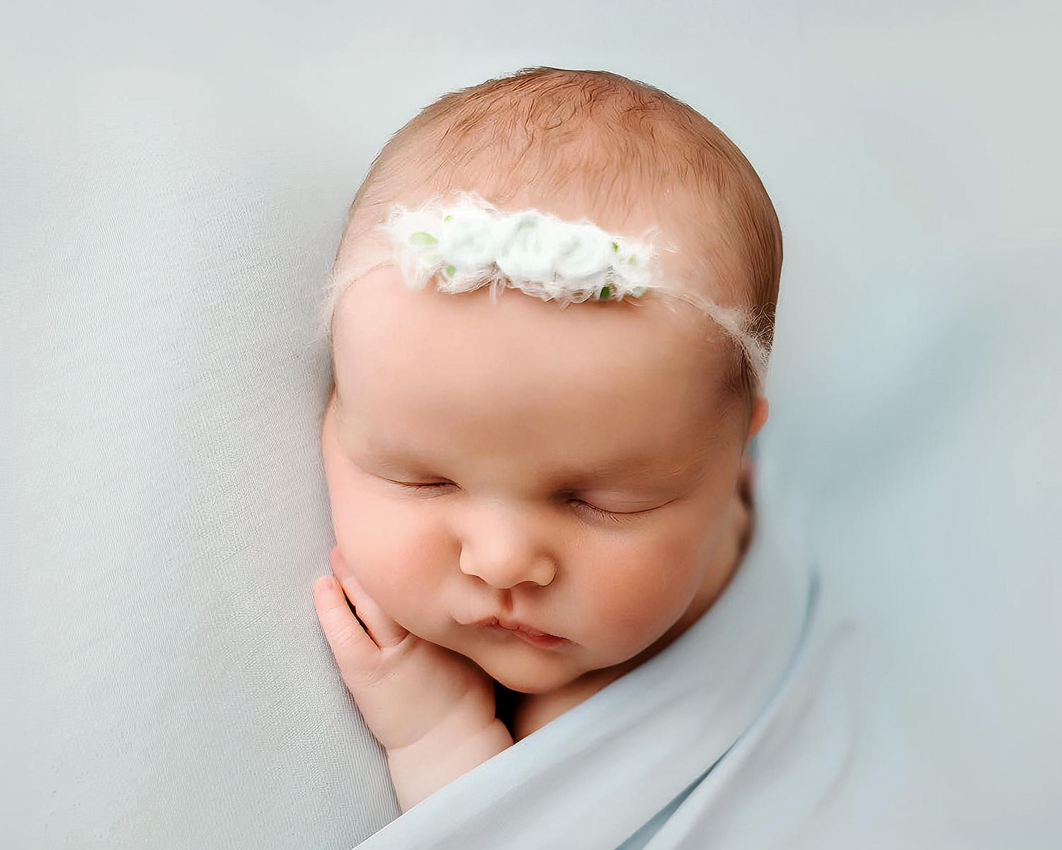 Sleeping newborn wearing a white floral headband wrapped in a soft blue wrap