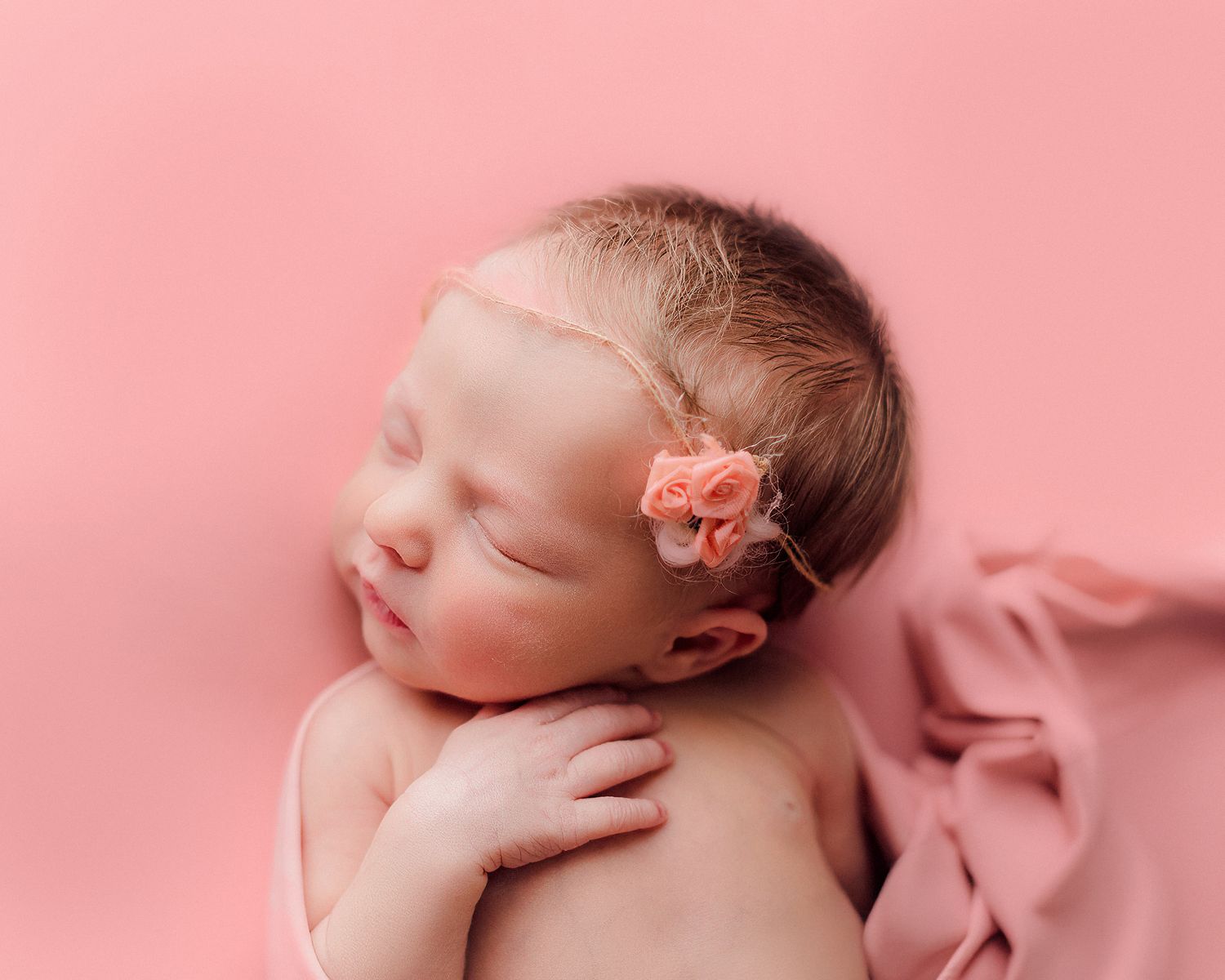 Sleeping newborn wrapped in pink fabric with a floral headband on a pink background