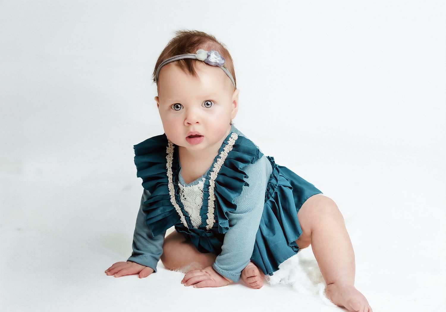 Baby sitting on a light blue background in a teal ruffled outfit, bright studio portrait