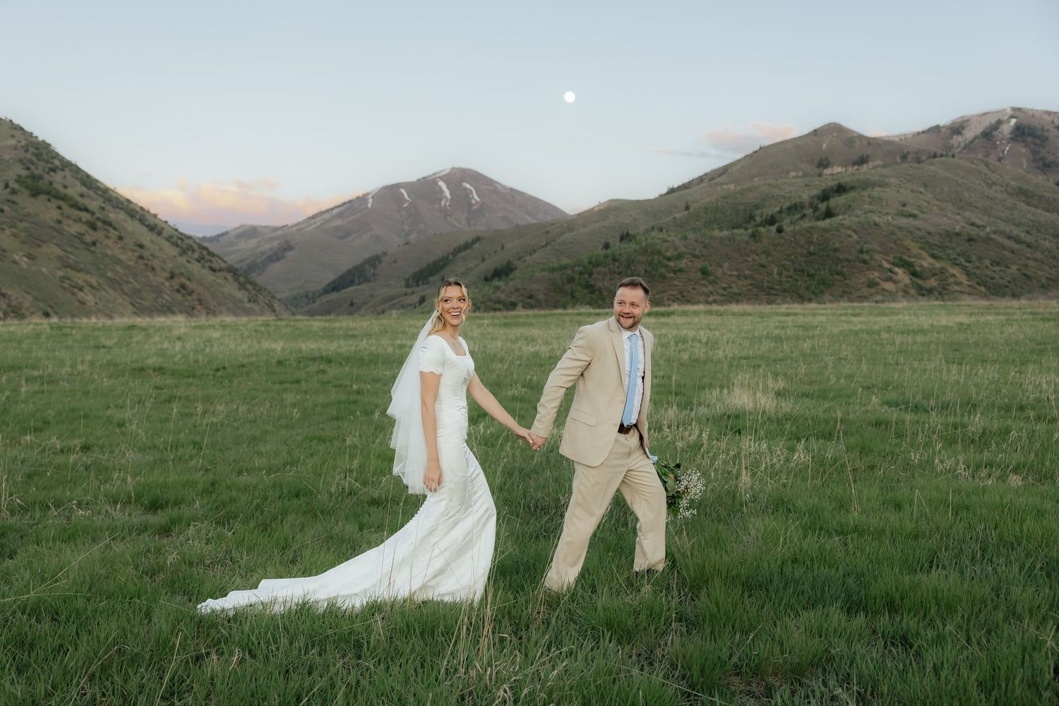 Couple walking holding hands in green field after sunset showcasing pink clouds and moon in Utah mountains.
