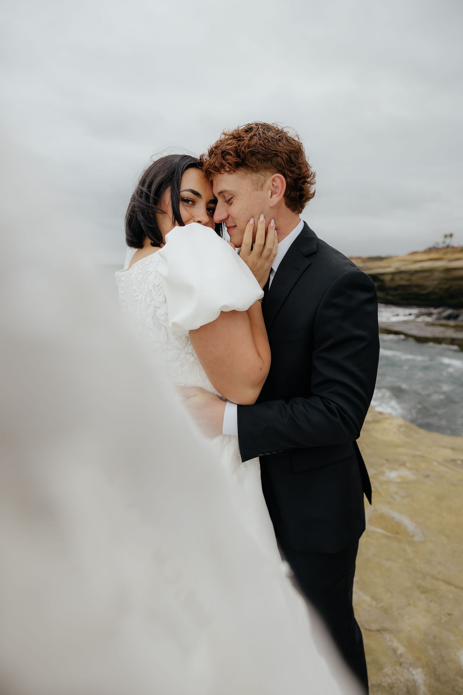 Bride and groom embracing on the rocky coastline during a destination wedding session in San Diego.