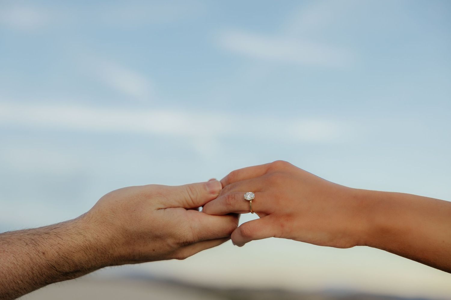 Artistic photo of couple holding hands showing wedding ring in front of a beautiful sunset sky.