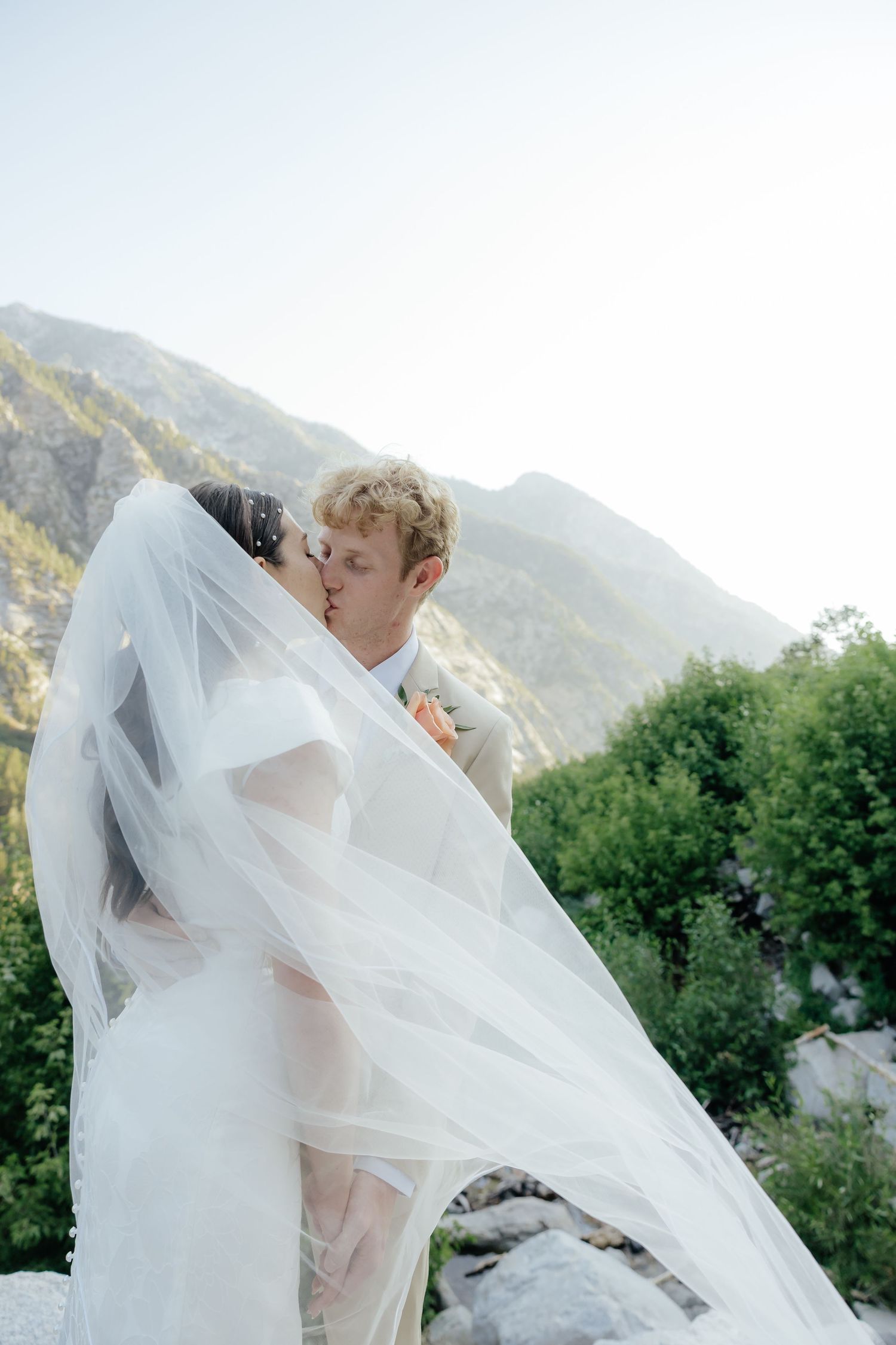 Couple kissing overlooking the Utah Mountains in the summertime showcasing bride's veil and wedding dress.