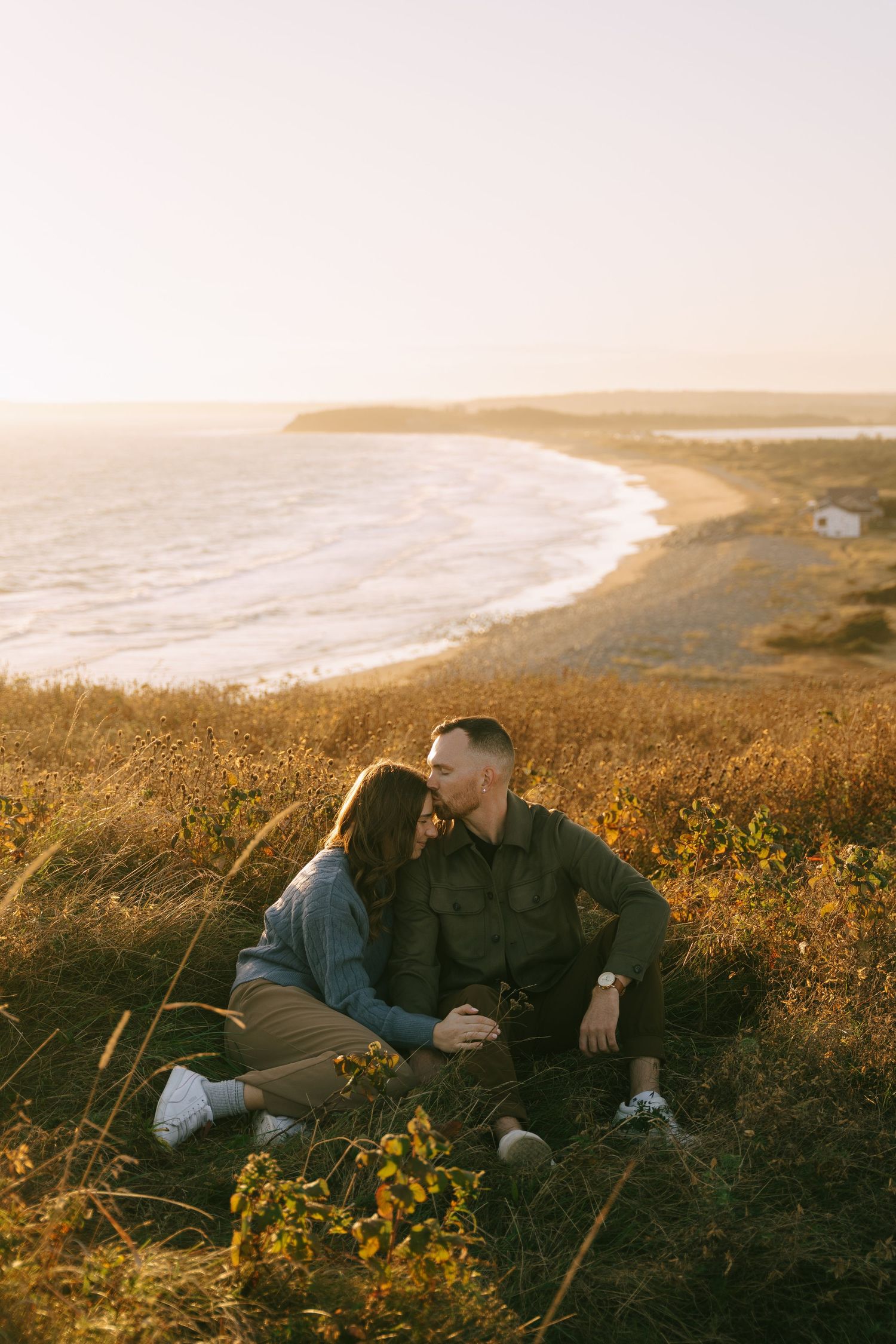 Two people sit together on a grassy coastal cliff overlooking the ocean during golden hour sunset in Halifax, Nova Scotia.