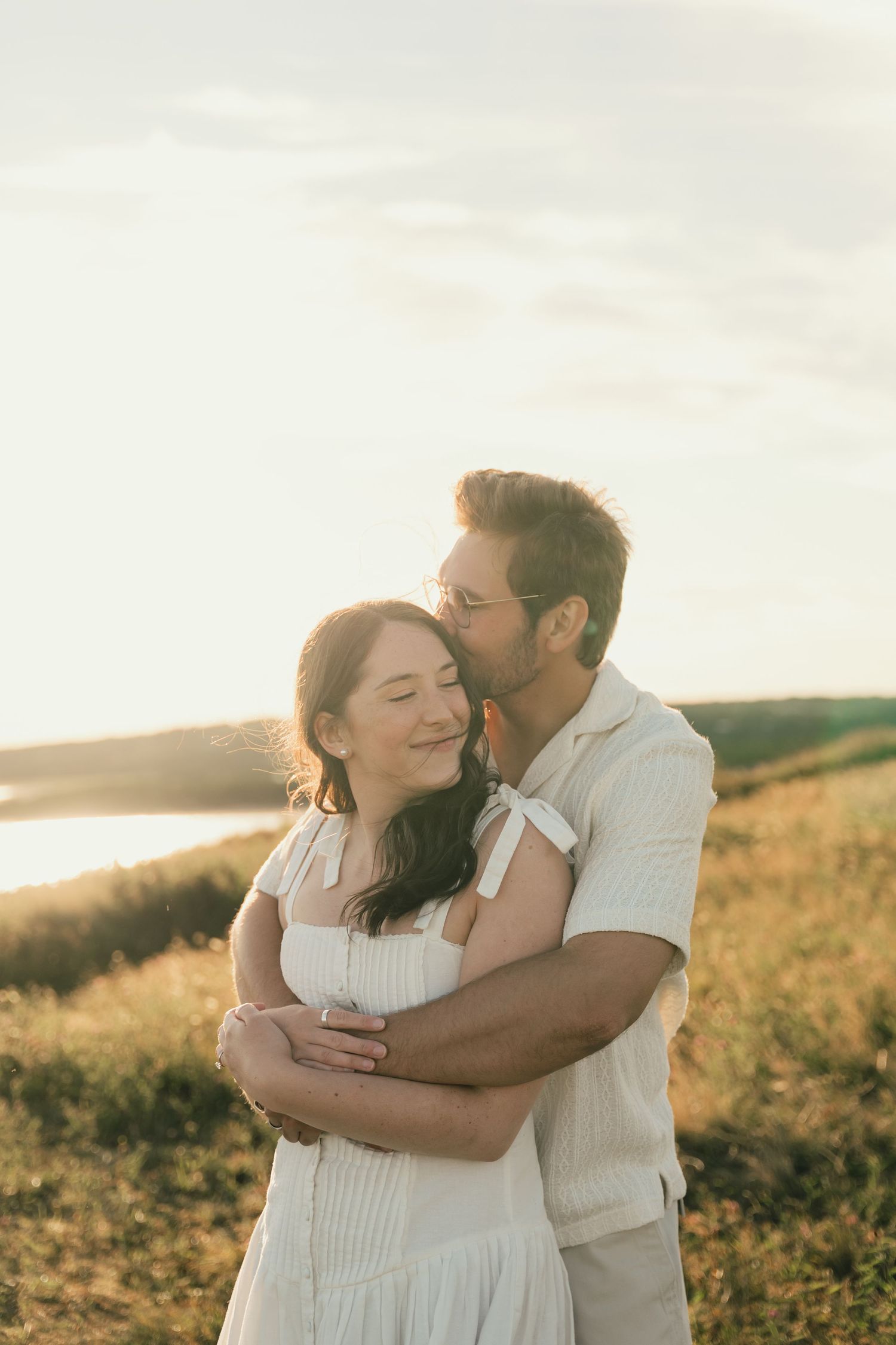 A tender moment captured between a couple wearing white as they embrace in a sunlit field in Halifax, Nova Scotia.