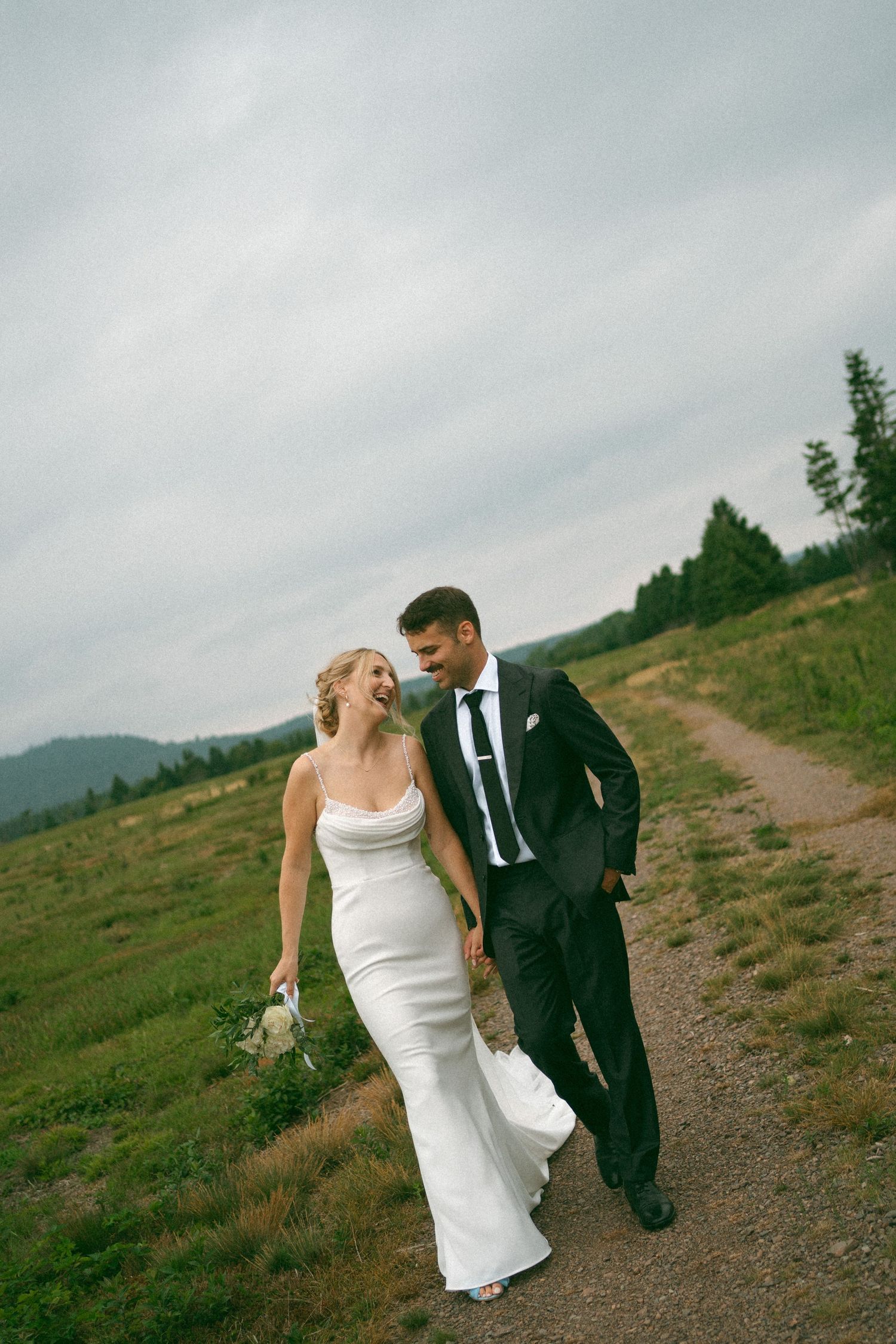 A couple in wedding attire walk along a scenic mountain trail in Nova Scotia under cloudy skies.