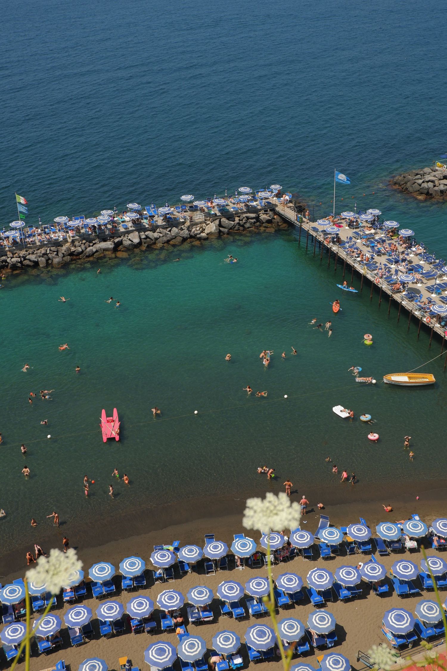 Aerial view of a beach club in Italy with blue umbrellas and swimmers enjoying the turquoise Mediterranean waters.