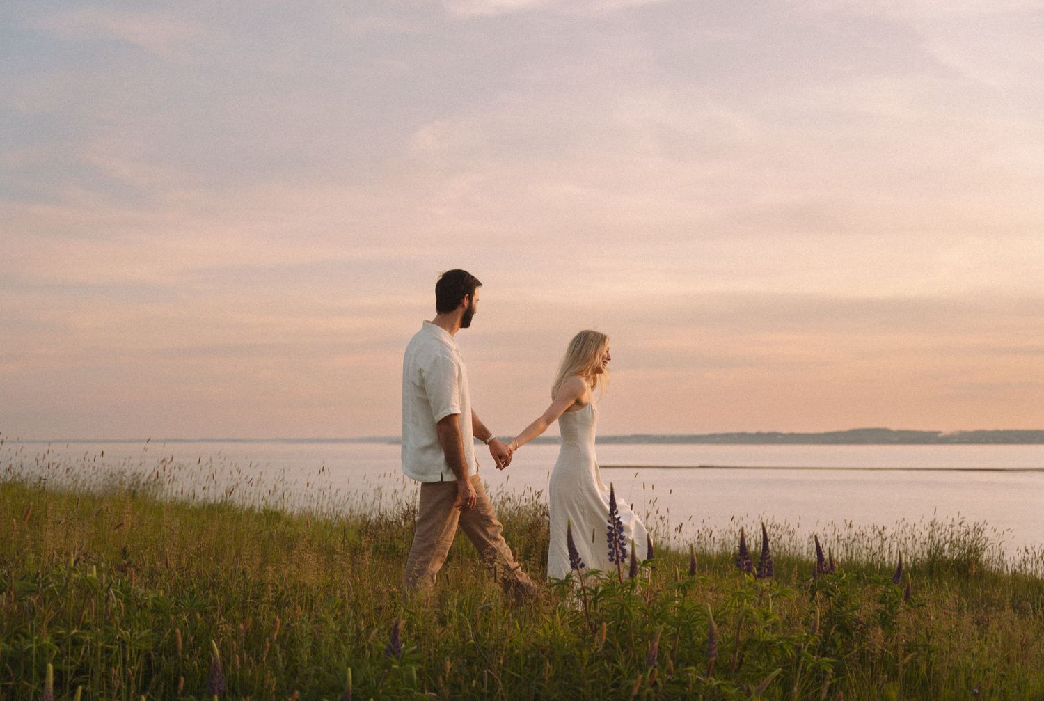 A couple walks hand in hand through tall grass along a serene waterfront in Halifax, Nova Scotia during a peaceful sunset.