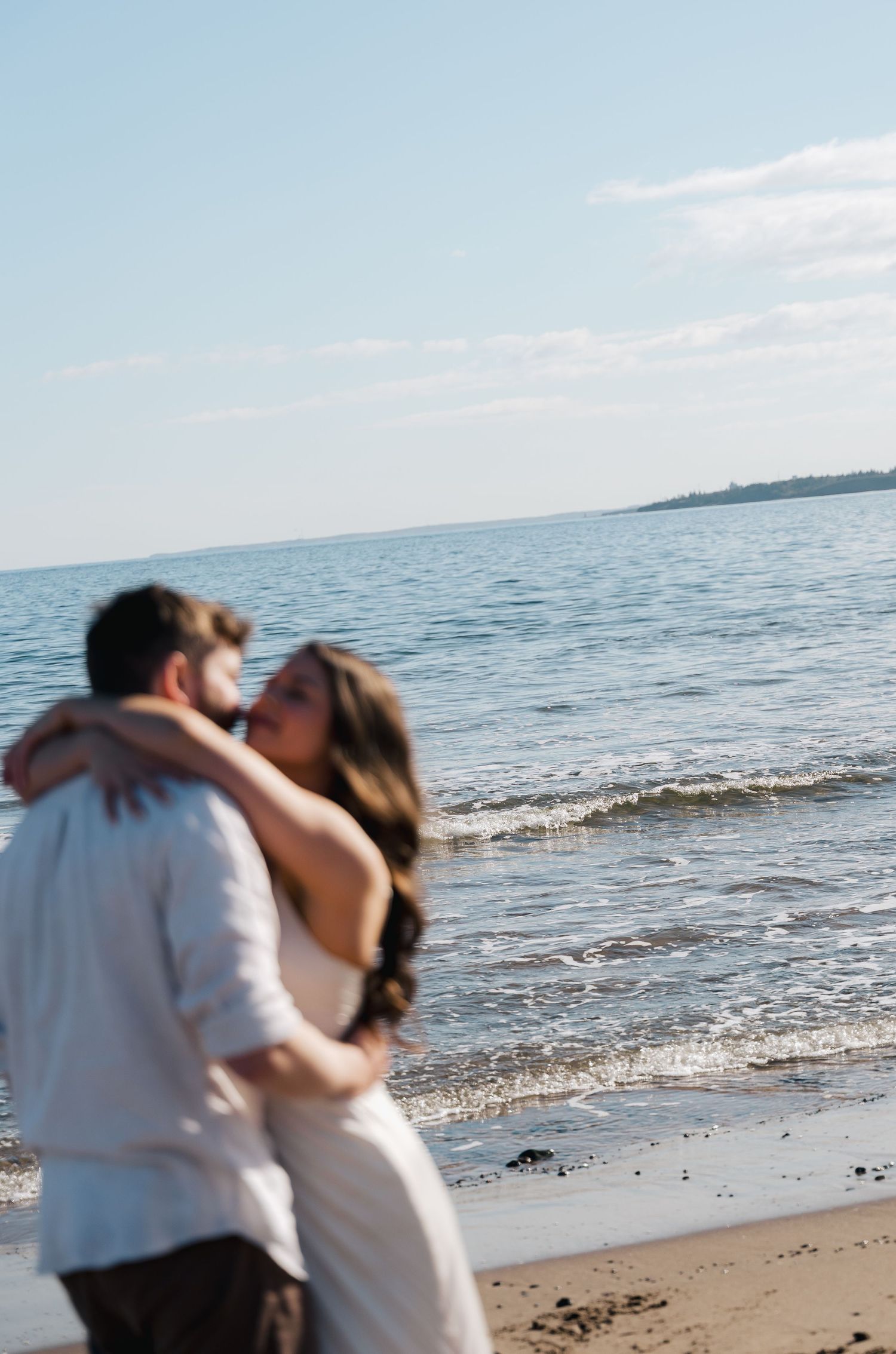 A romantic couple shares a tender embrace on a serene beach in Halifax, Nova Scotia with gentle waves and blue sky in the background.