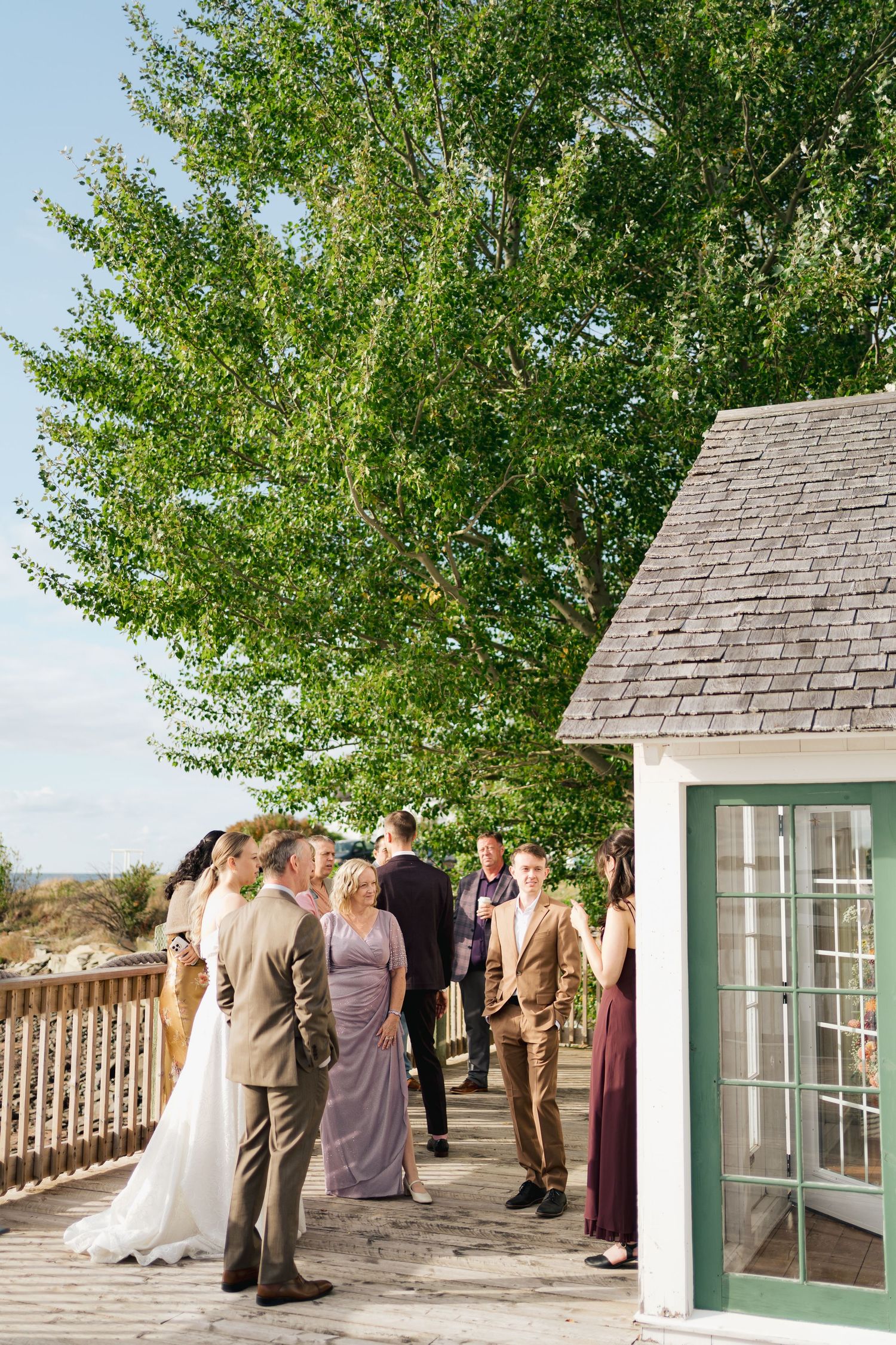 Wedding party gathered on an outdoor patio with green trees and white railings in the background in Nova Scotia.
