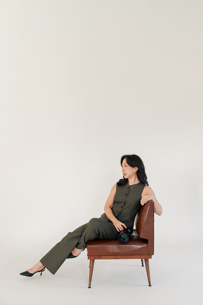 Woman in olive green jumpsuit lounges elegantly on vintage brown leather chair against white backdrop.