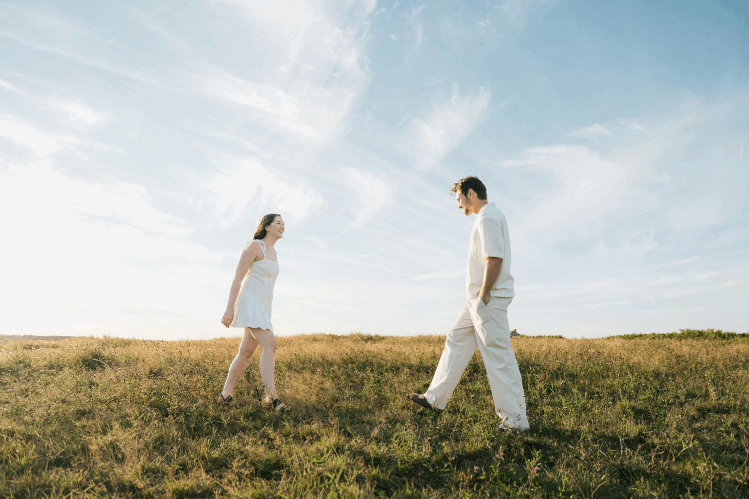 Two people in white outfits walk through a grassy hilltop meadow in Halifax, Nova Scotia against a bright blue summer sky.