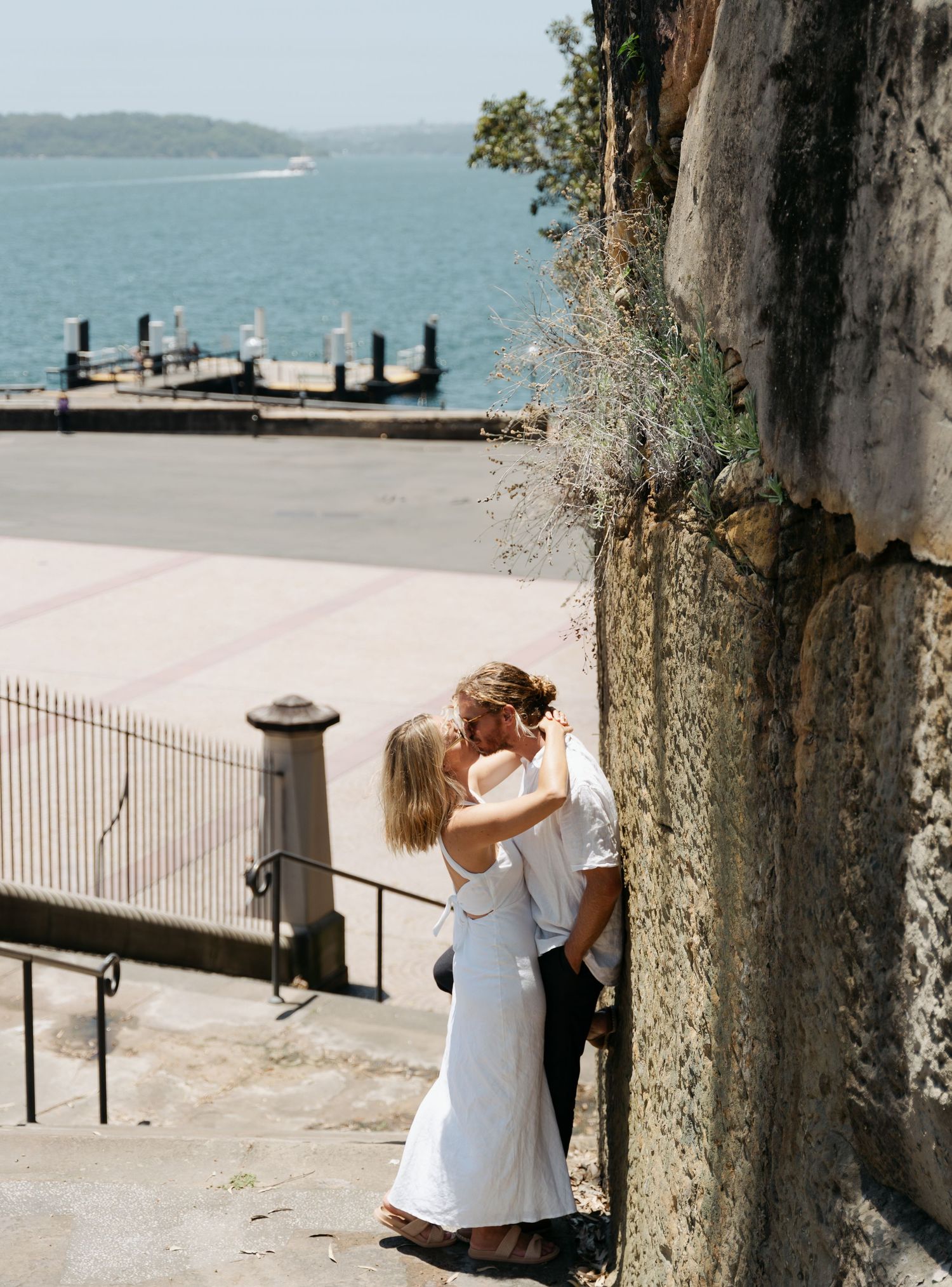 A couple in white outfits shares romantic moments against stone pillars by a waterfront setting in Sydney.