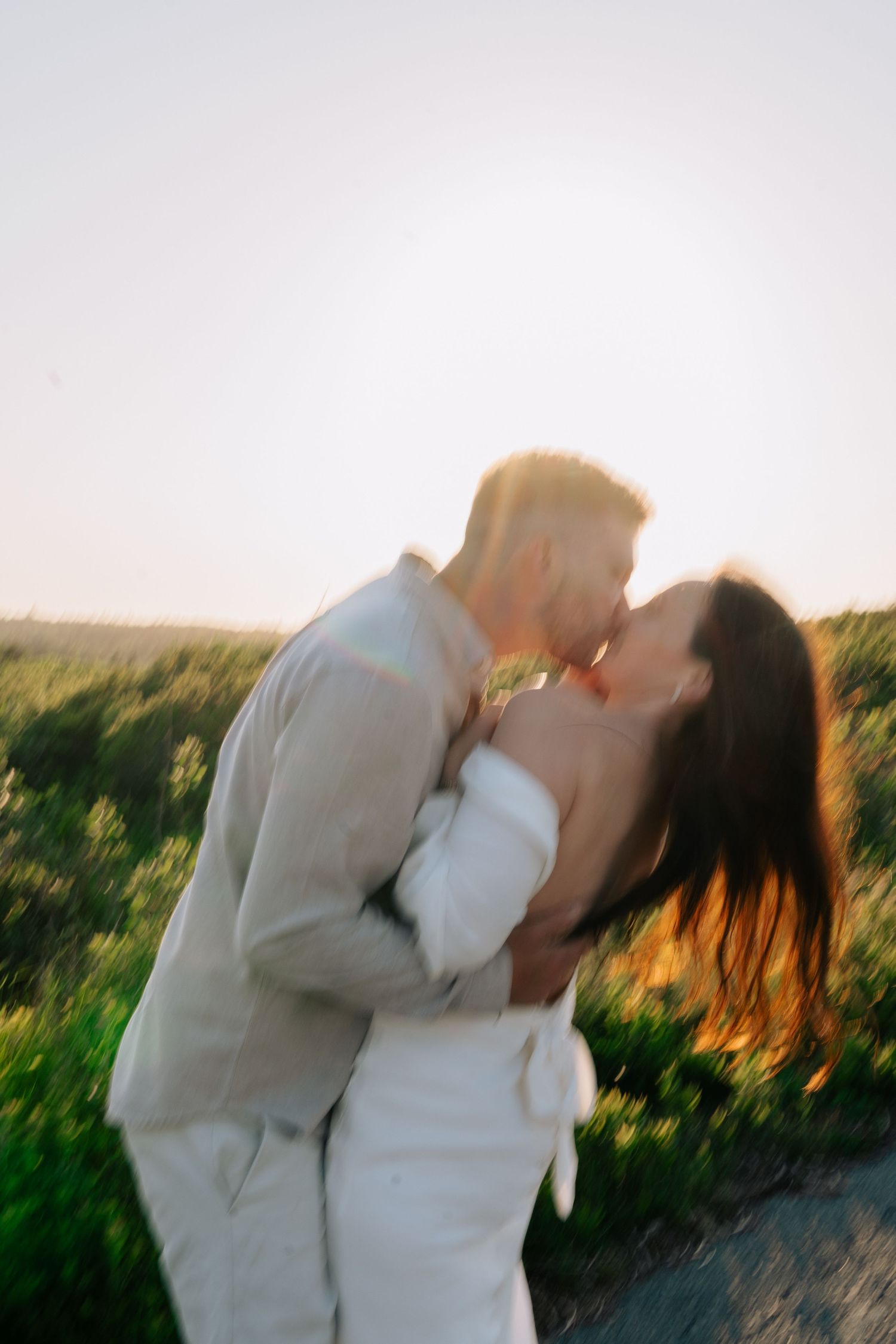 A couple in white embraces and shares a romantic kiss at sunset in a grassy field in Halifax, Nova Scotia.