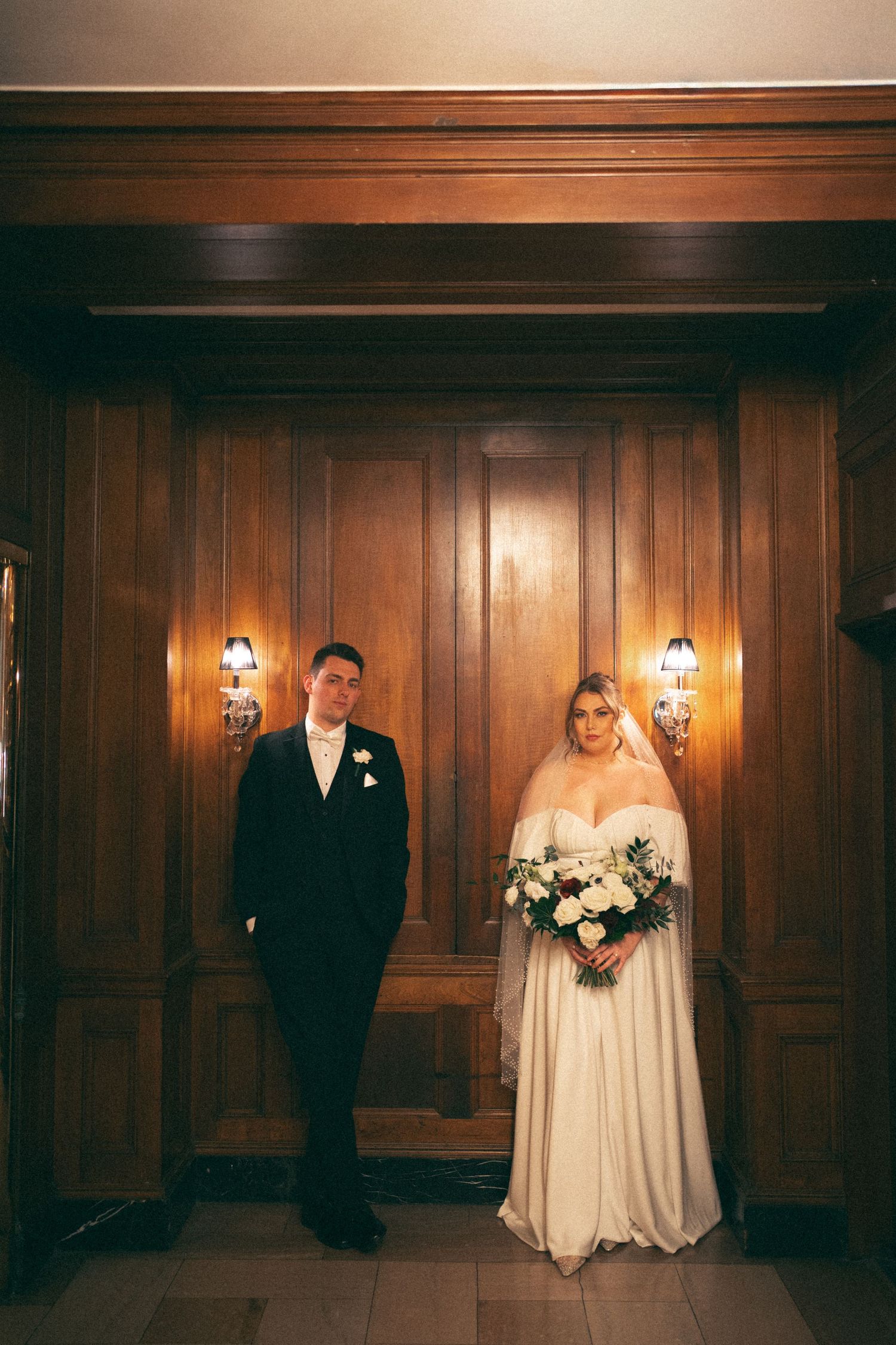 Wedding couple poses against dark wood-paneled wall with warm lighting and elegant sconces at Lord Nelson Hotel, Halifax, Nova Scotia.