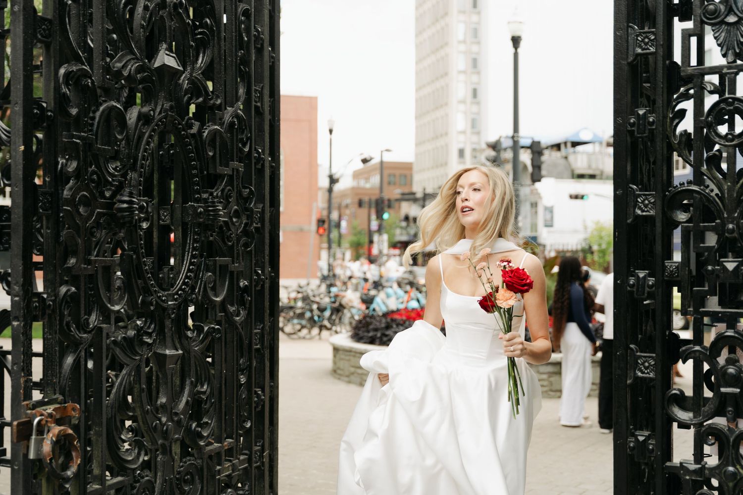 A person in a white wedding dress holds red flowers while standing near ornate black iron gates in an urban setting in Halifax, Nova Scotia.