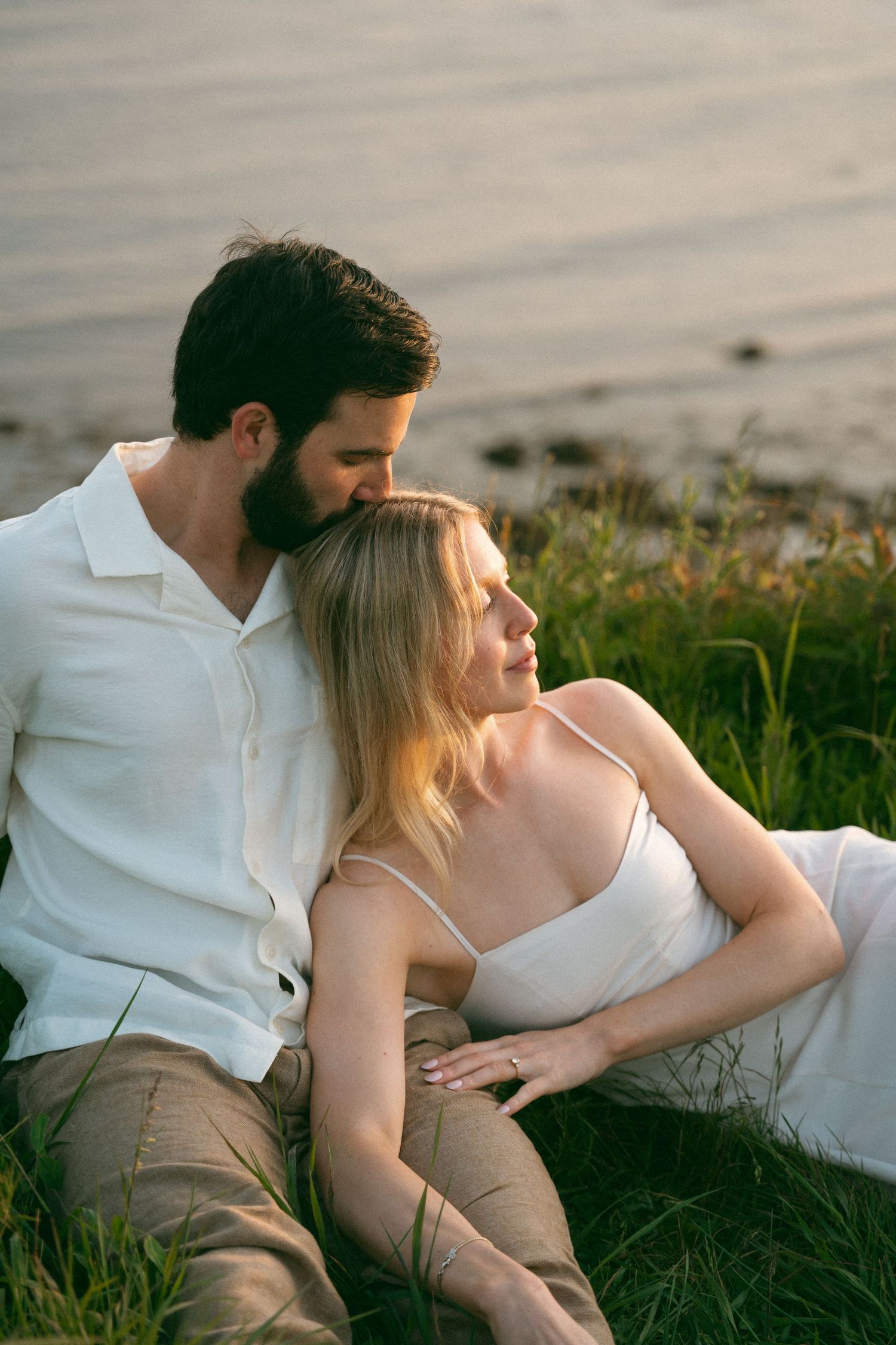 A couple sits together in a grassy meadow located in Lawrencetown Halifax, Nova Scotia during golden hour, dressed in white and beige tones.