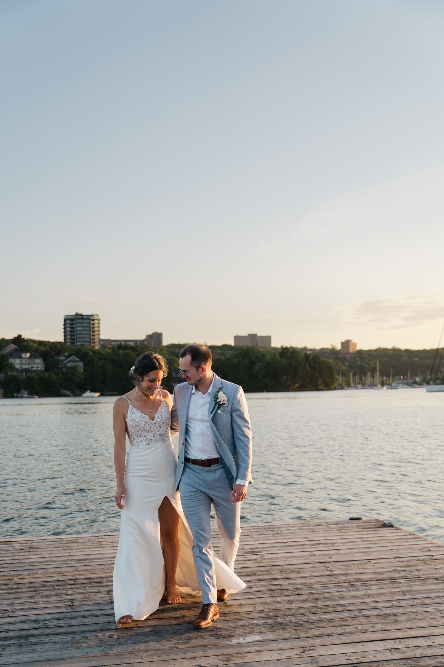 A couple sits together on a wooden dock at sunset overlooking a calm lake with city buildings in the background in Halifax, Nova Scotia.