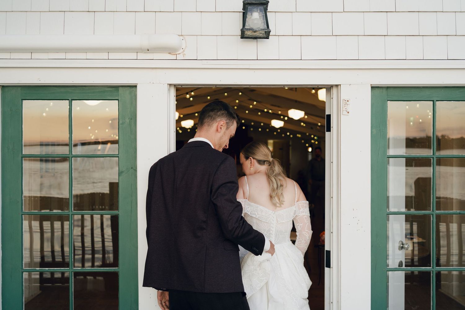 A couple in wedding attire stands in a doorway decorated with string lights at sunset in Nova Scotia.