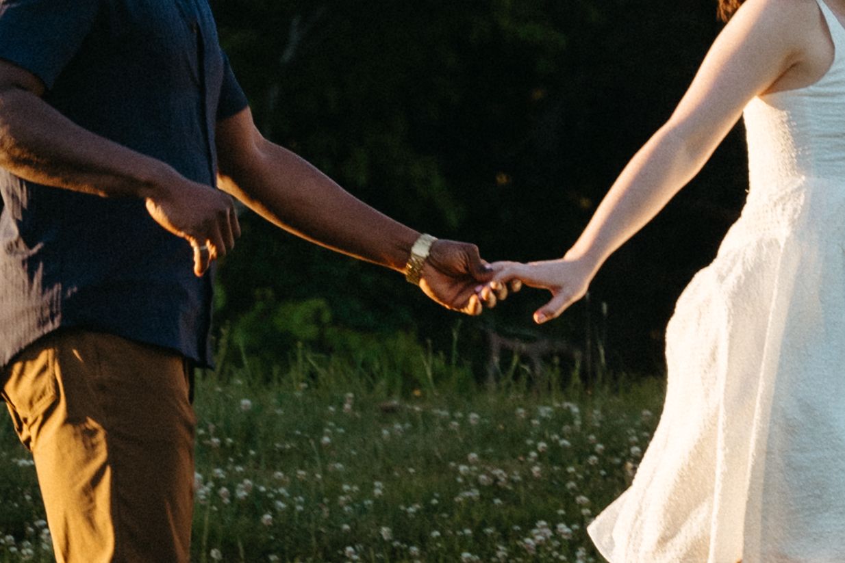 Two people holding hands in a sunlit meadow with wildflowers, one wearing a white dress and another in navy and yellow in Halifax, NS.