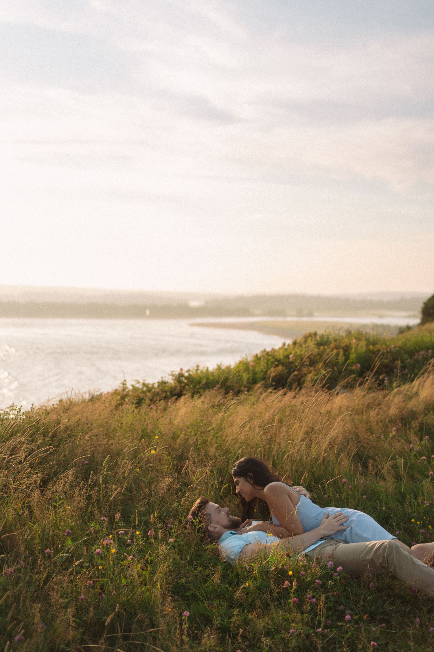 A couple lies in tall grass overlooking a scenic coastal view at sunset in Lawrencetown, Halifax, Nova Scotia.