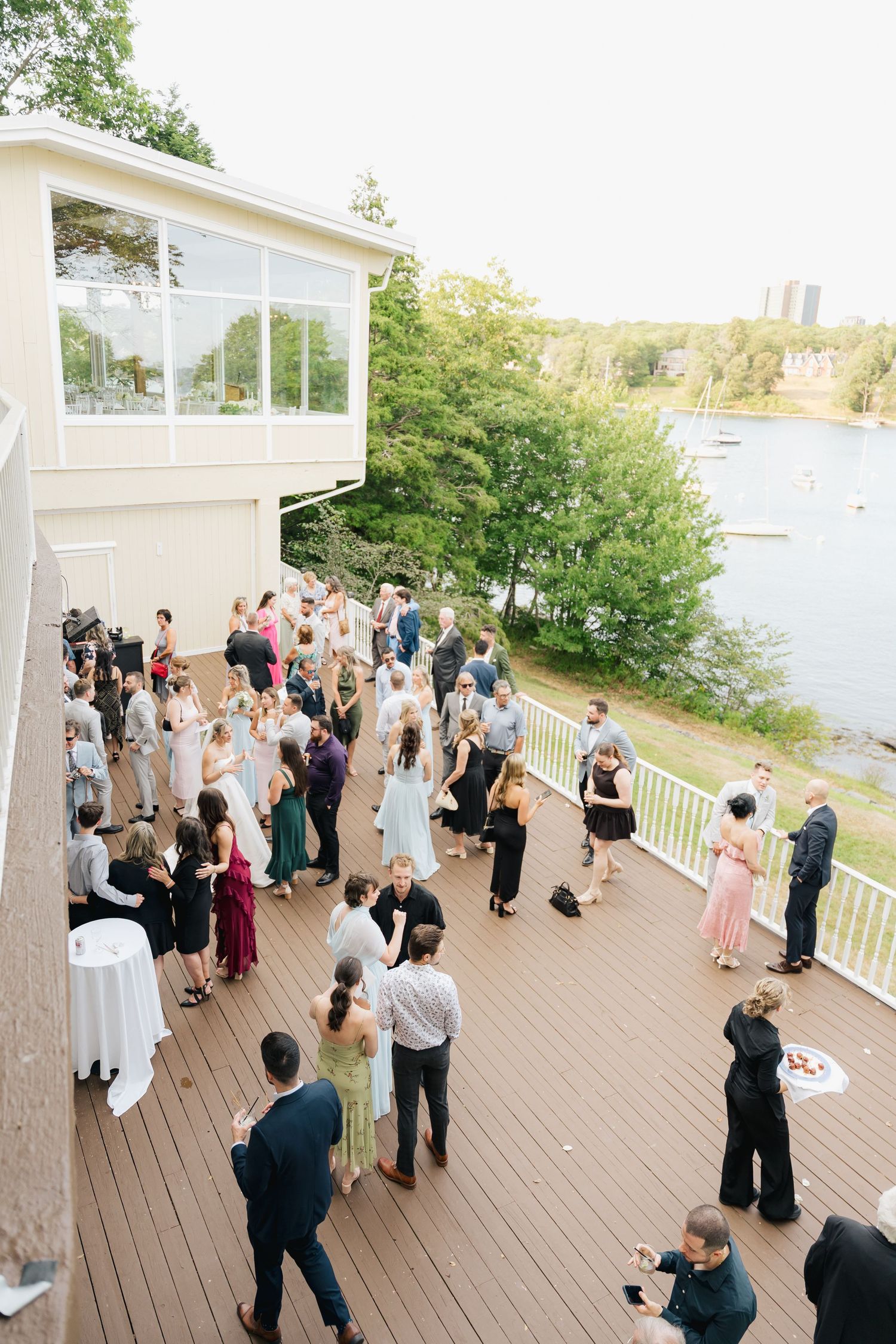 Wedding guests gather on a waterfront deck during an outdoor reception overlooking a scenic bay in Halifax, Nova Scotia.