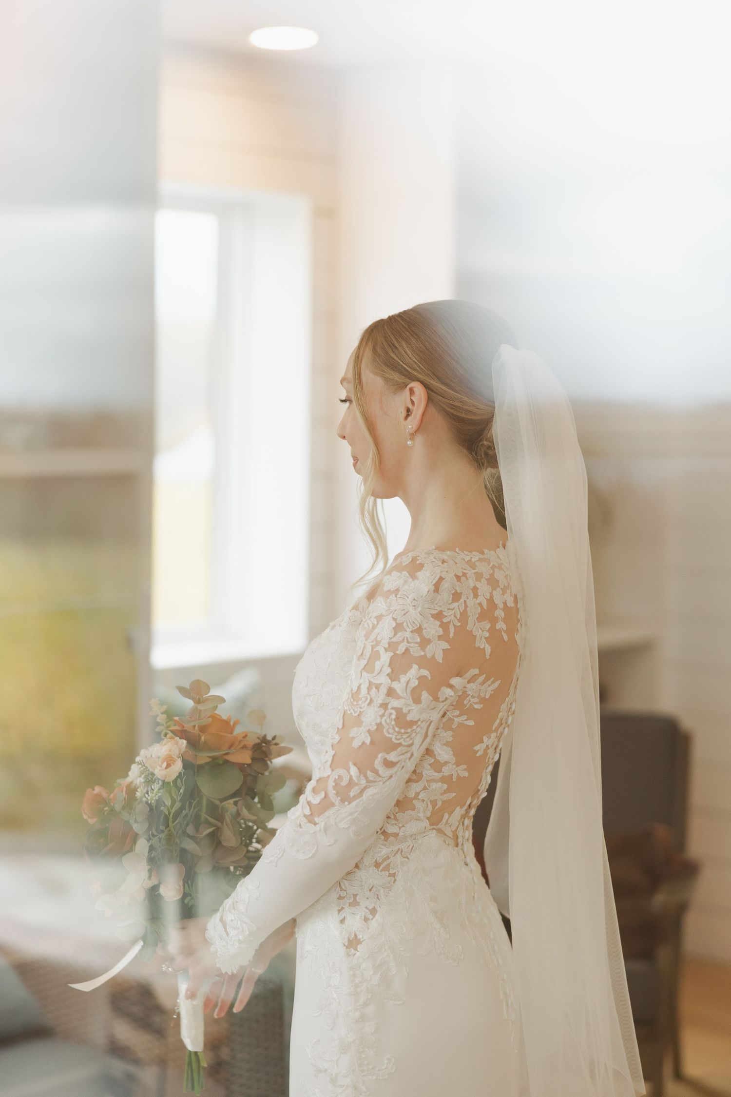 A bride in a delicate lace wedding dress stands by a window in soft natural light at Lightfoot & Wolfville.