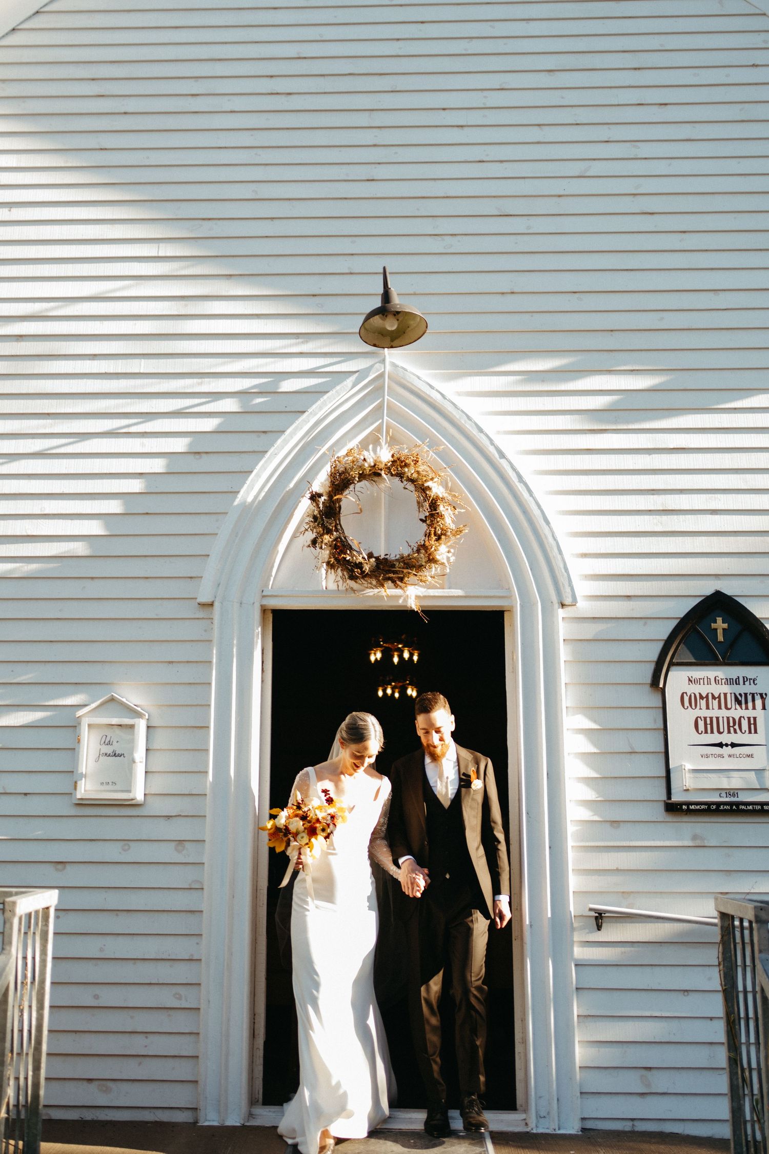 A wedding couple exits through white arched church doorways in warm sunlight in Nova Scotia.