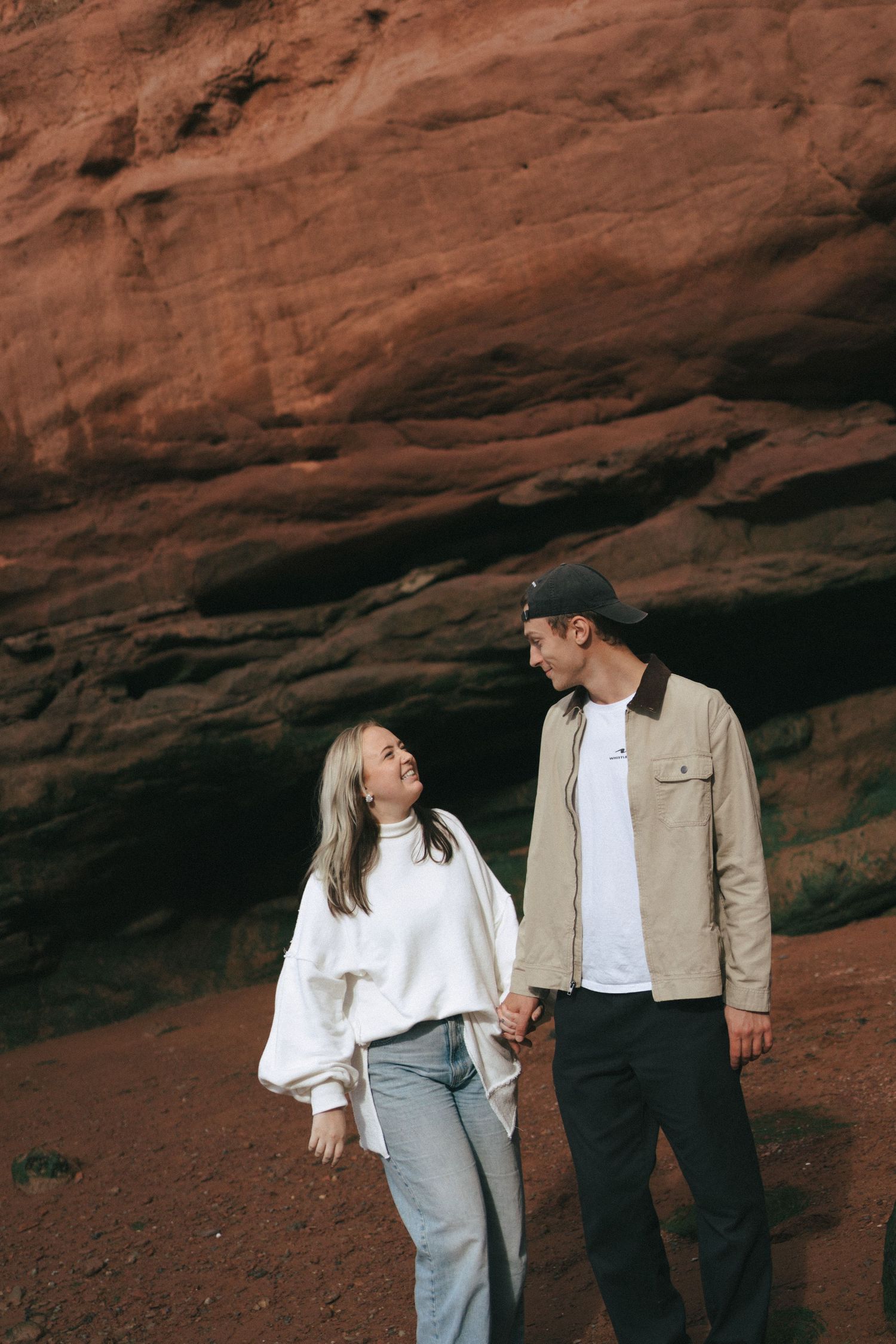 A romantic couple in casual attire embraces against a dramatic red rock formation in a desert landscape in Nova Scotia.