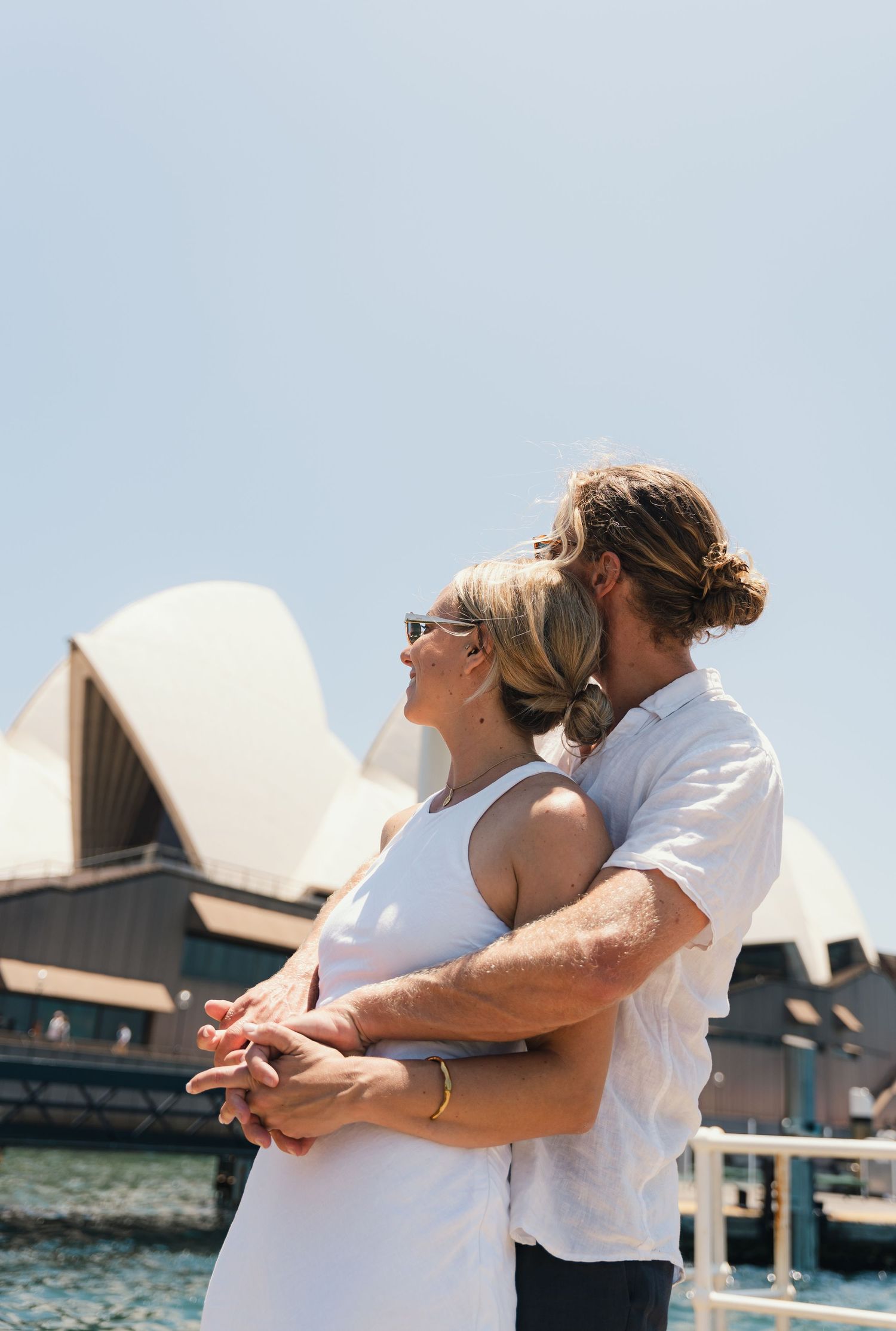 A couple embraces while enjoying the view of Sydney Opera House on a sunny day at Sydney Harbour.
