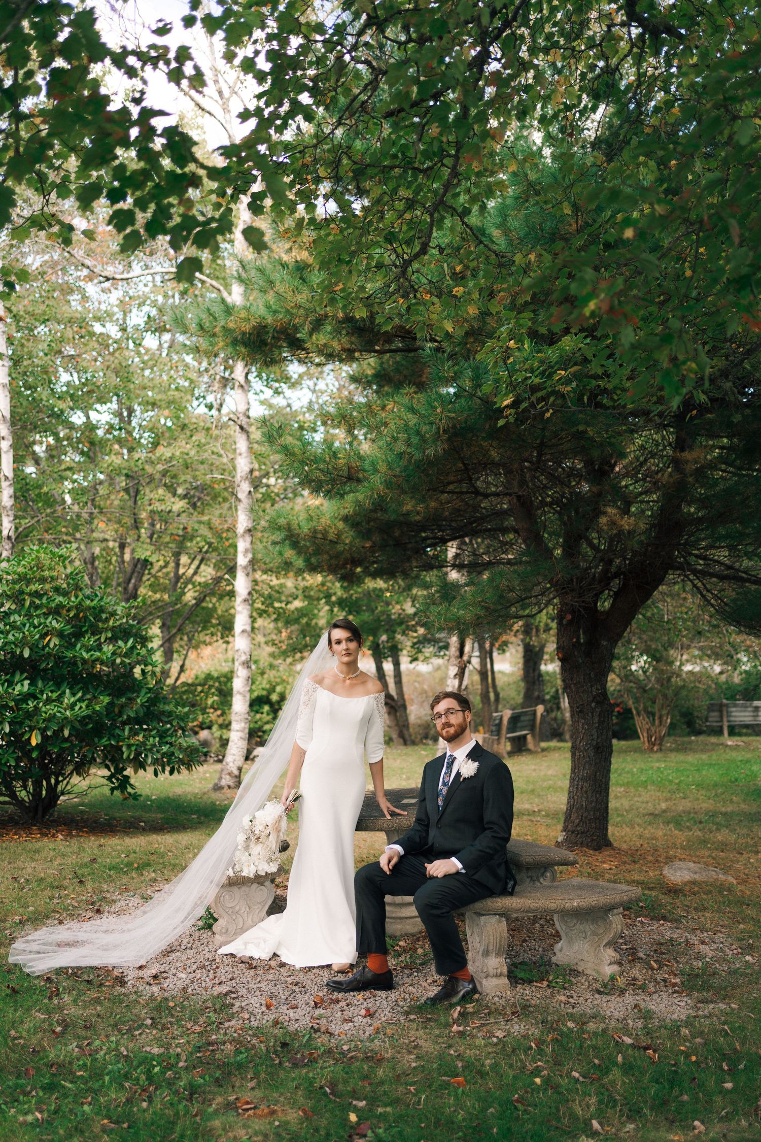 Wedding couple sits together on fallen leaves under green trees in a romantic outdoor forest setting in Halifax, Nova Scotia.