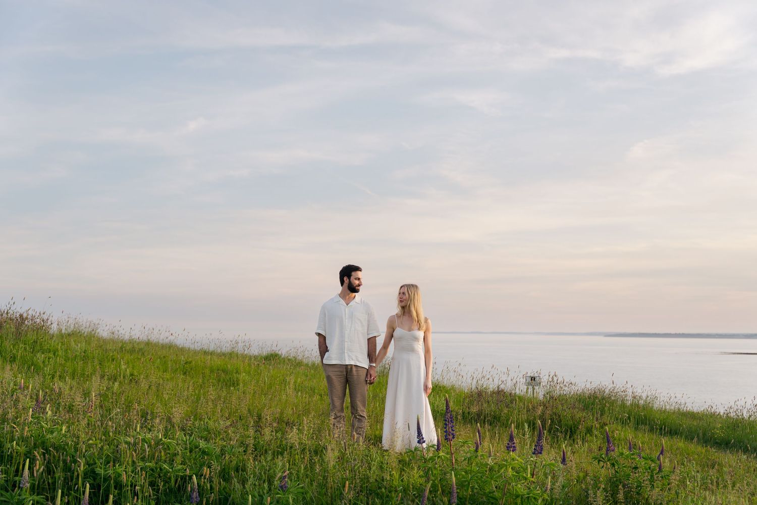 Couple in white outfits walking through a serene coastal meadow at sunset with ocean views in the background in Halifax, Nova Scotia.