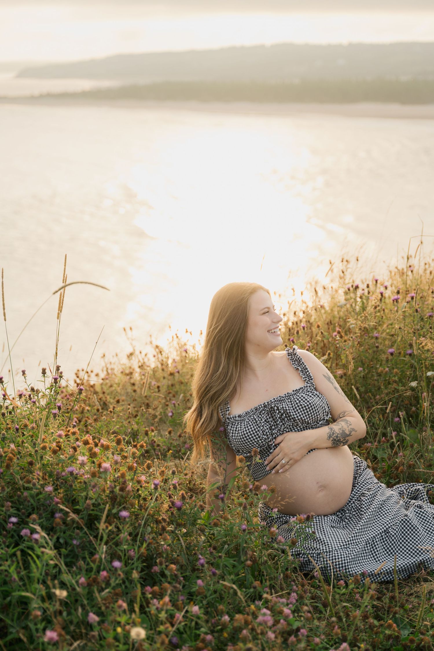 An expectant mother in a gingham dress sits among wildflowers at golden hour on a maternity photoshoot in Lawrencetown, Halifax Nova Scotia.