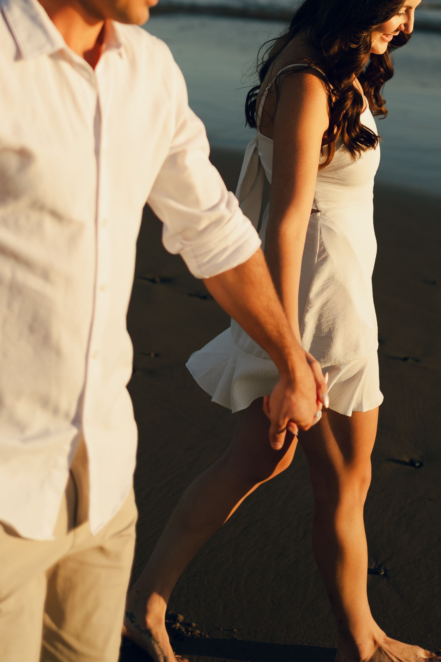 A couple in white and black outfits holds hands while walking on the beach in Halifax, Nova Scotia during sunset.