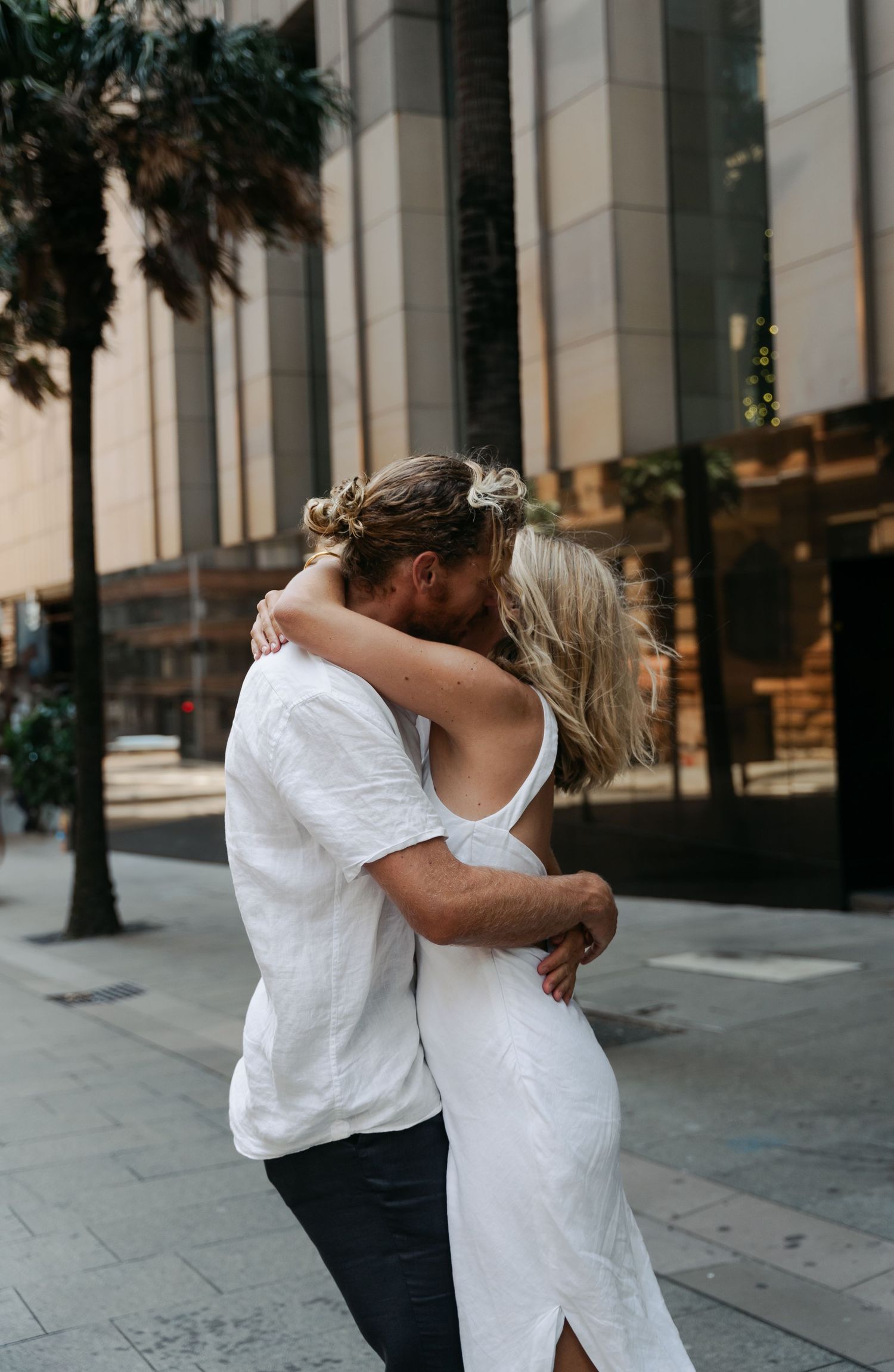 A couple in white clothing embraces and dances together on a city sidewalk near modern buildings.