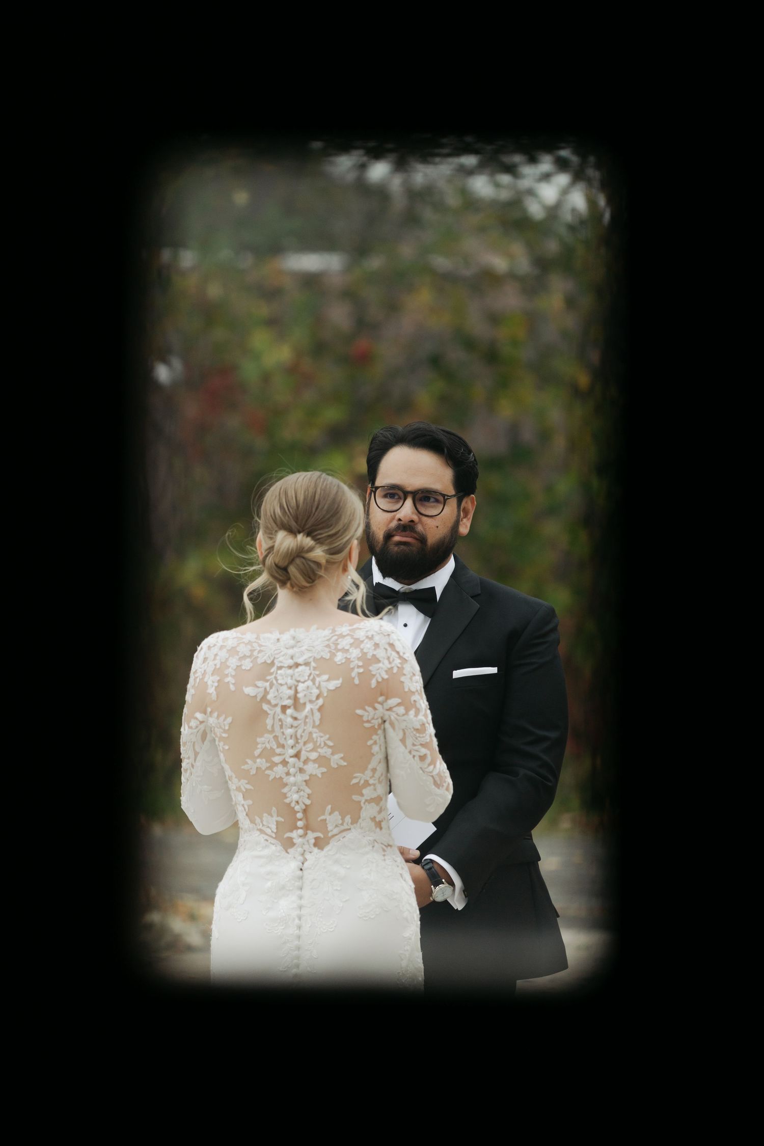 A romantic wedding couple embraces outdoors with the bride wearing an elegant lace-detailed dress with sheer back details.