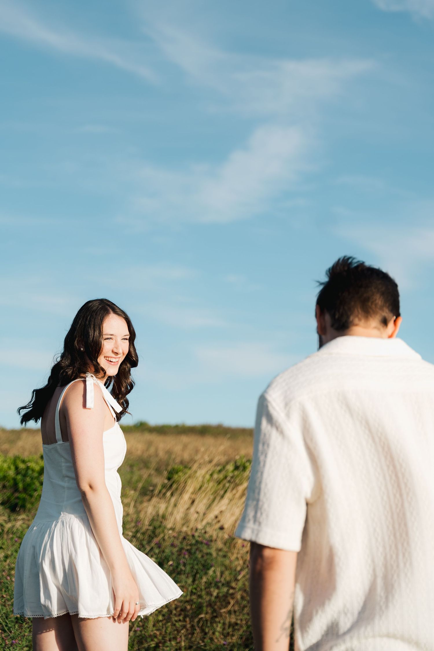 A couple in white outfits share a romantic moment in a sunny field in Halifax, Nova Scotia with tall grass and blue skies.