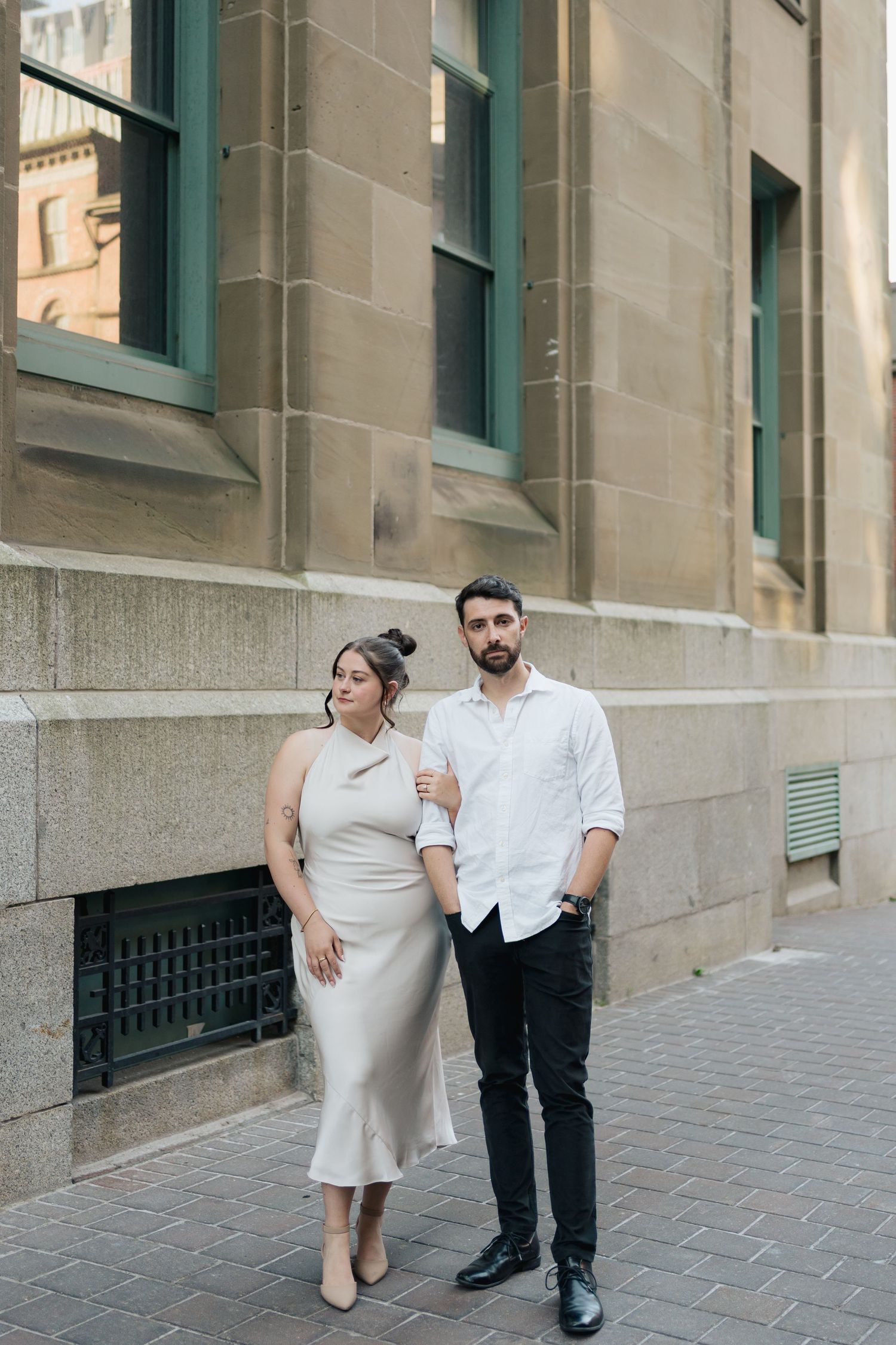 A couple in coordinated white outfits poses together against a stone building with teal-trimmed windows in Halifax, Nova Scotia.