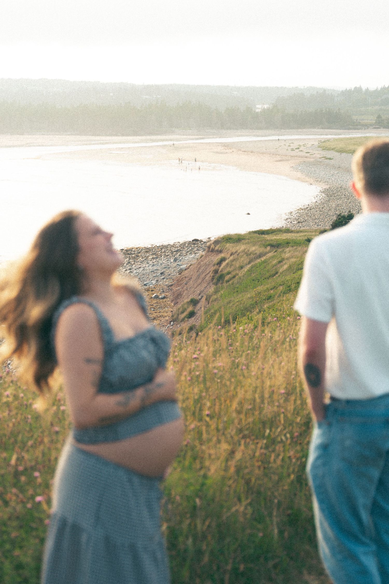 A couple embraces while standing in tall grass on a coastal hillside during golden hour in Lawrencetown, Halifax, Nova Scotia.