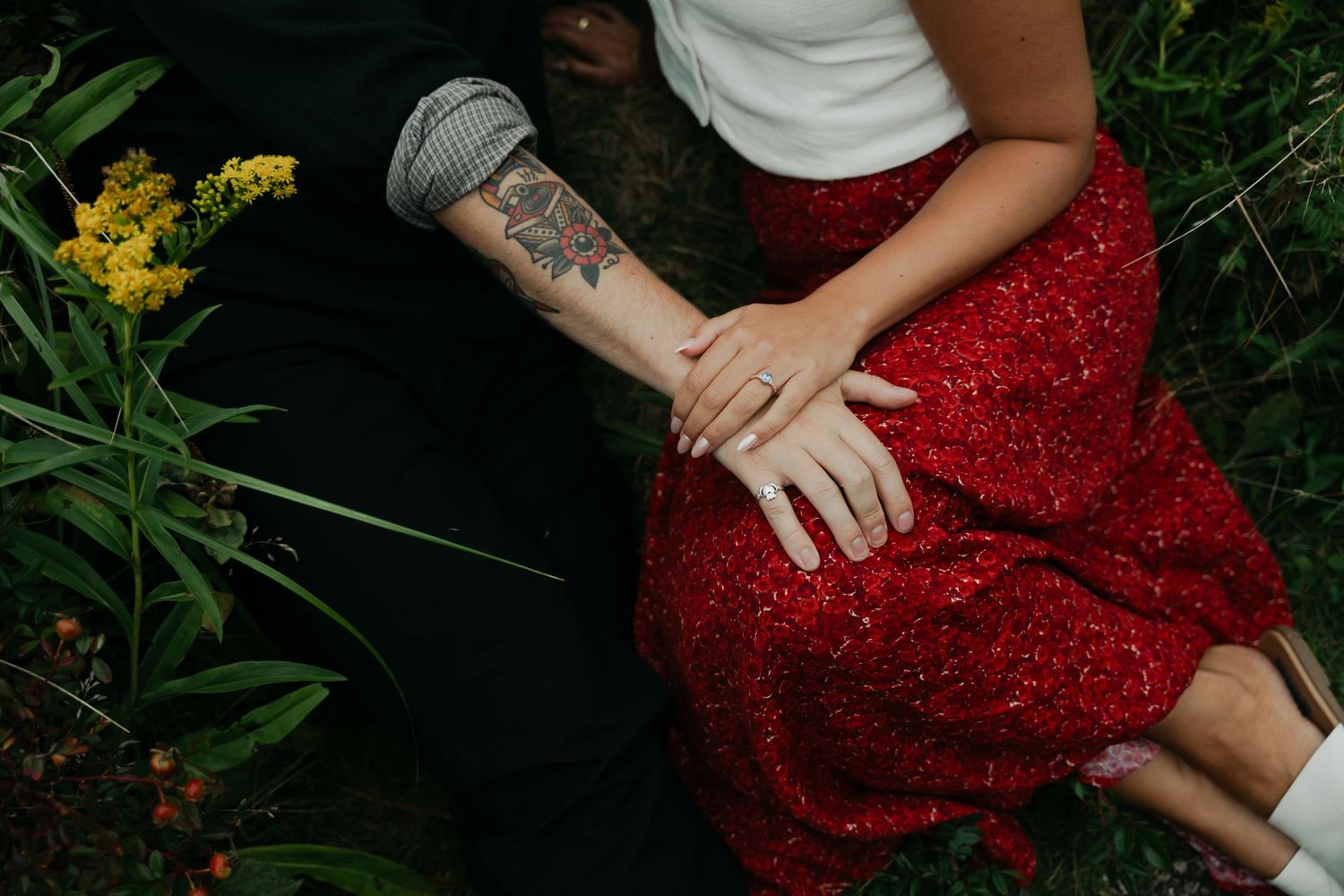 A couple sitting in grass in Halifax, Nova Scotia with a tattoo visible on their forearm near yellow wildflowers.