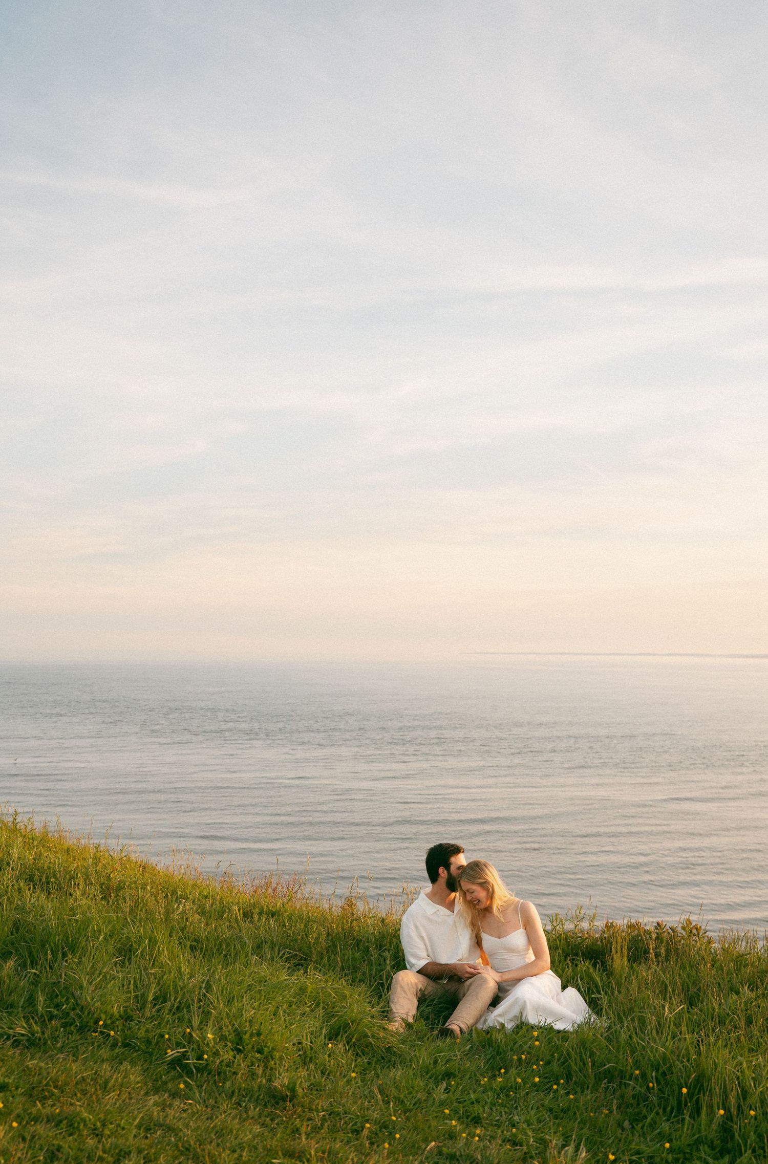 A couple sits together on a grassy coastal cliff overlooking a serene ocean at sunset in Lawrencetown, Halifax, Nova Scotia.