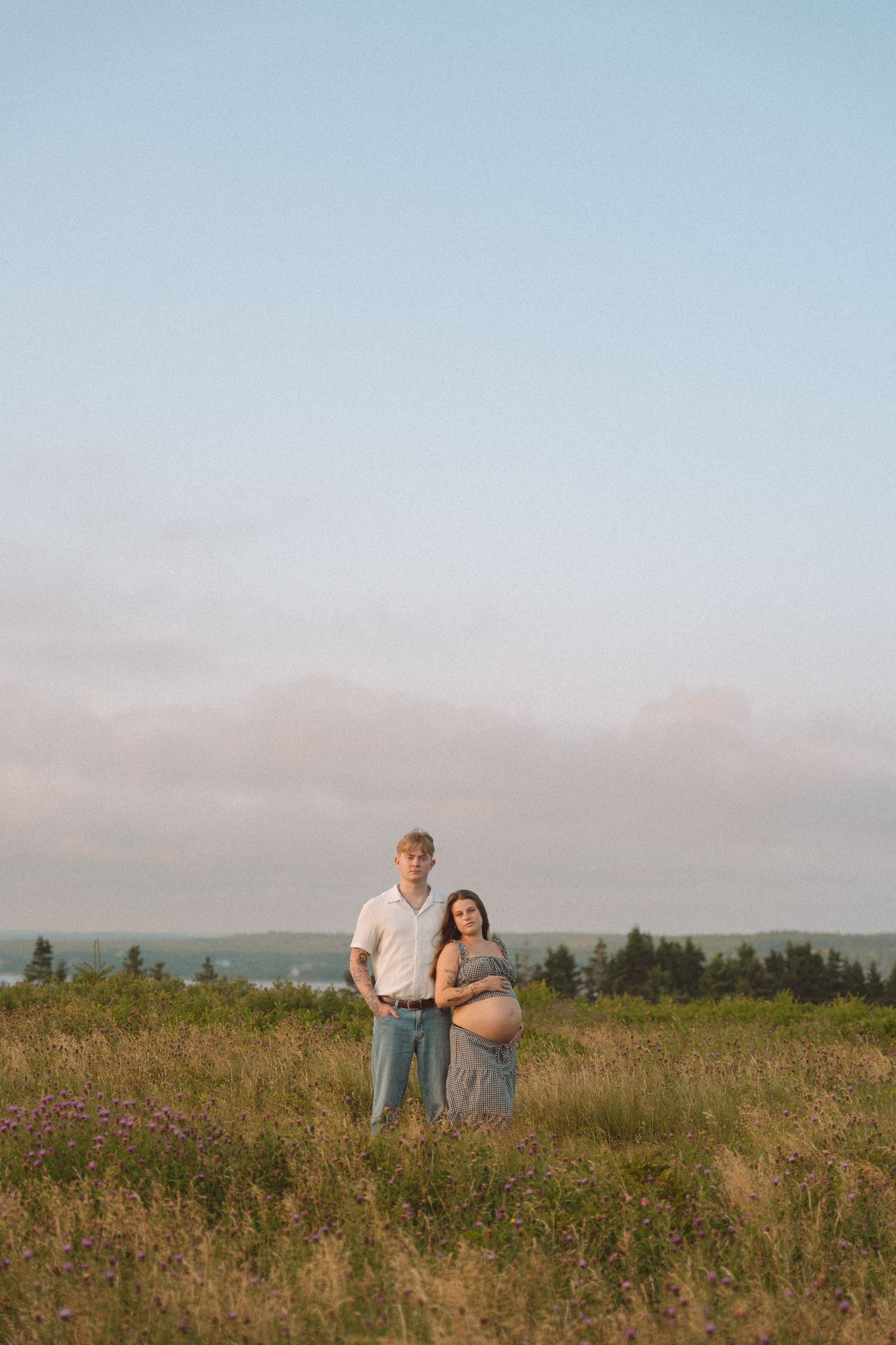 A couple stands together in a scenic wildflower meadow at sunset with mountains in the distance in Lawrencetown, Halifax, Nova Scotia.