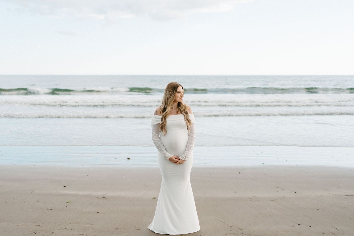 A pregnant woman in a flowing white dress stands on a misty beach with waves rolling in the background in Halifax, Nova Scotia.