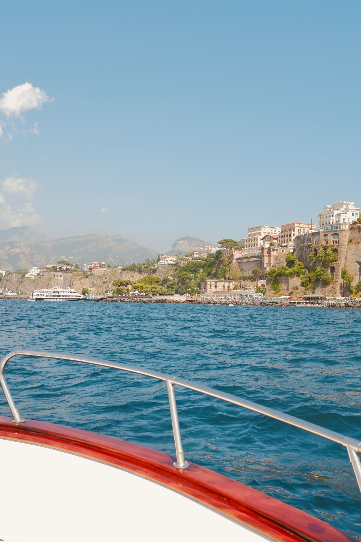 Panoramic view of a coastal Italian town from a boat with red railing showing blue waters and hillside buildings.