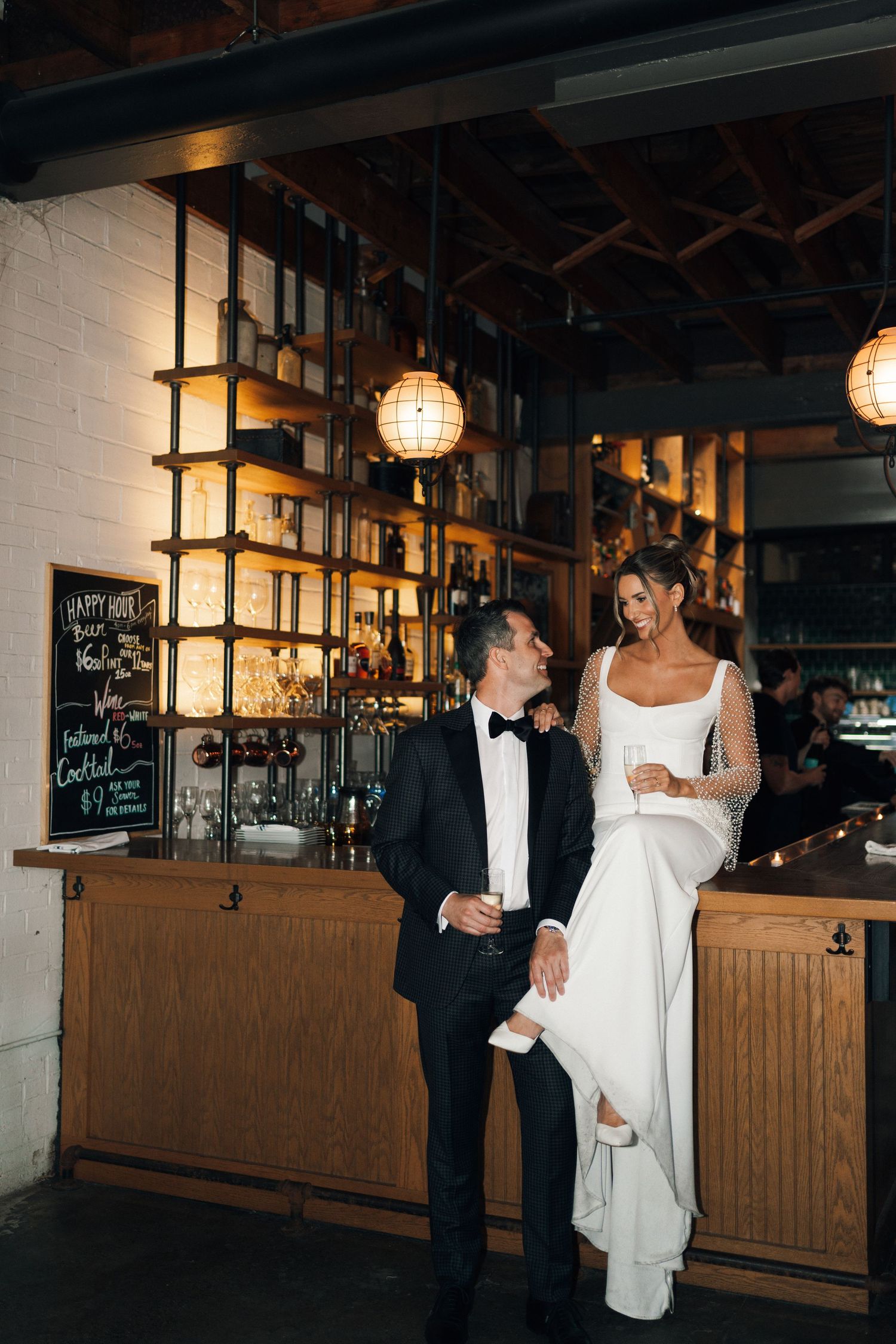 A couple shares a joyful moment at a rustic bar with wooden shelving and pendant lighting during their wedding celebration in Halifax, NS.