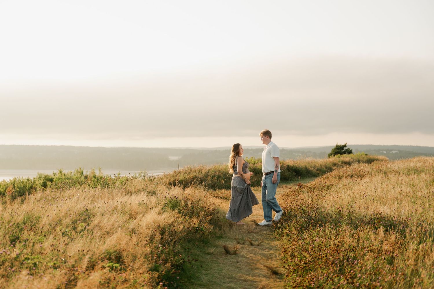 Two people sit together on a grassy hilltop overlooking a scenic coastal landscape in Halifax, Nova Scotia at golden hour.