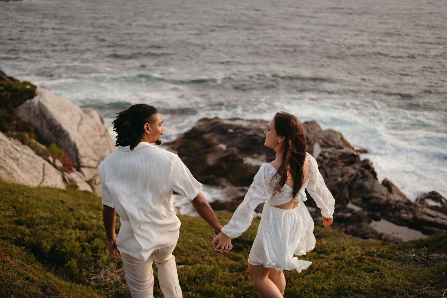 Couple in white outfits walking along rocky coastal cliffs at sunset with ocean waves crashing below in Halifax, Nova Scotia.
