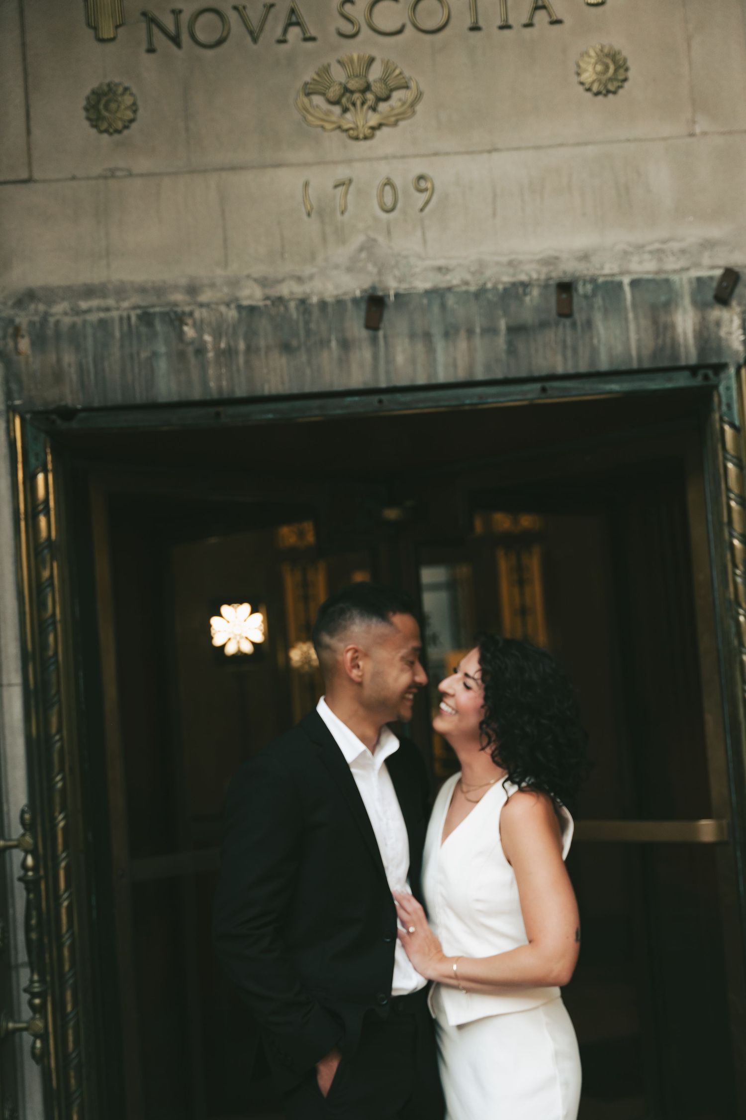 A couple in white outfits share an intimate moment in front of a rustic building entrance in downtown Halifax, Nova Scotia.