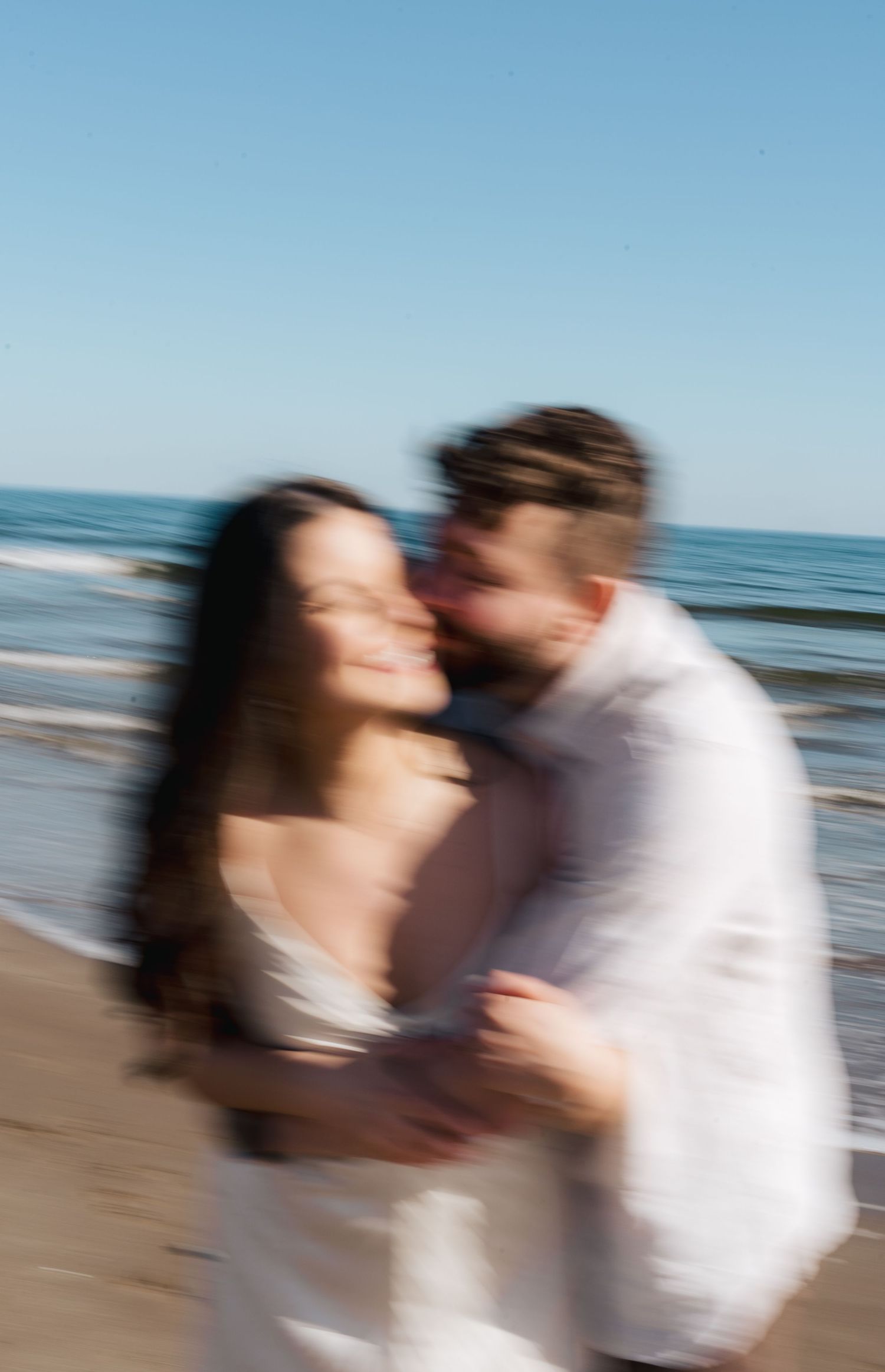 A blurred romantic moment captured on the beach in Halifax, Nova Scotia with ocean waves and blue sky in the background.