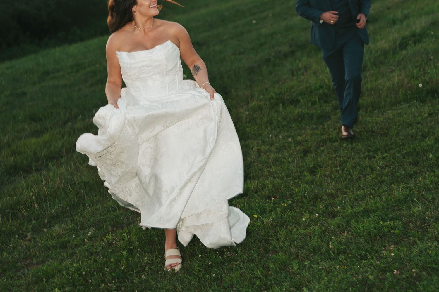 A bride in a flowing white wedding dress walks across a green lawn during an outdoor wedding in Halifax, Nova Scotia.